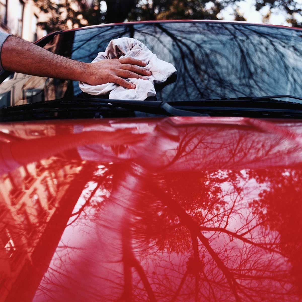 A hand wipes a white cloth across a shiny red car hood, reflecting tree branches and a building in a natural outdoor setting.