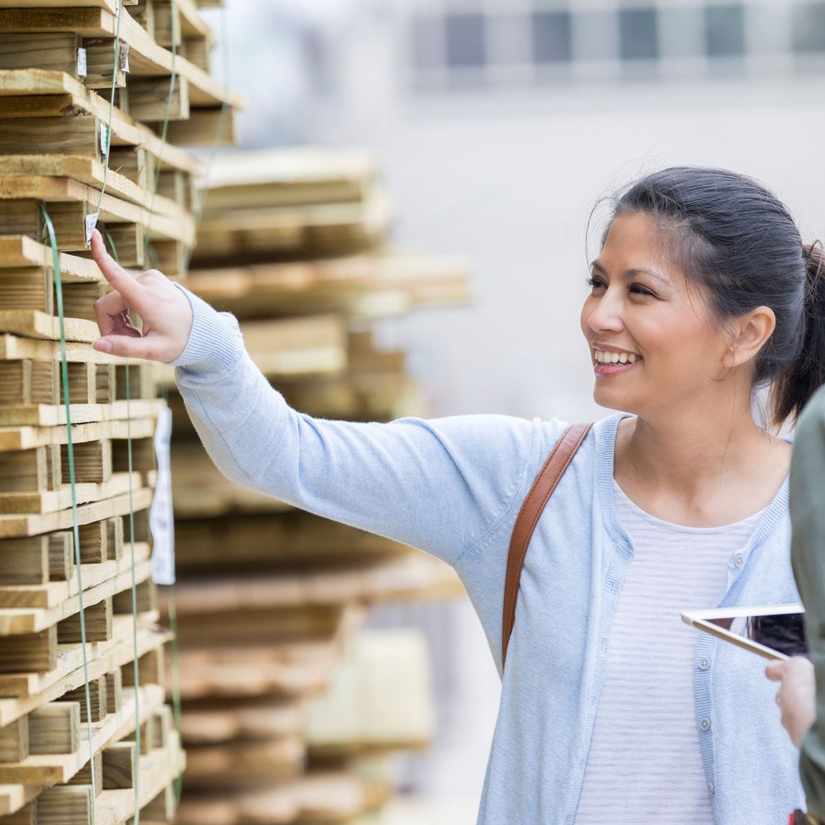 A woman points at items on stacked pallets, smiling while engaging with someone nearby, in an outdoor setting filled with wooden materials.