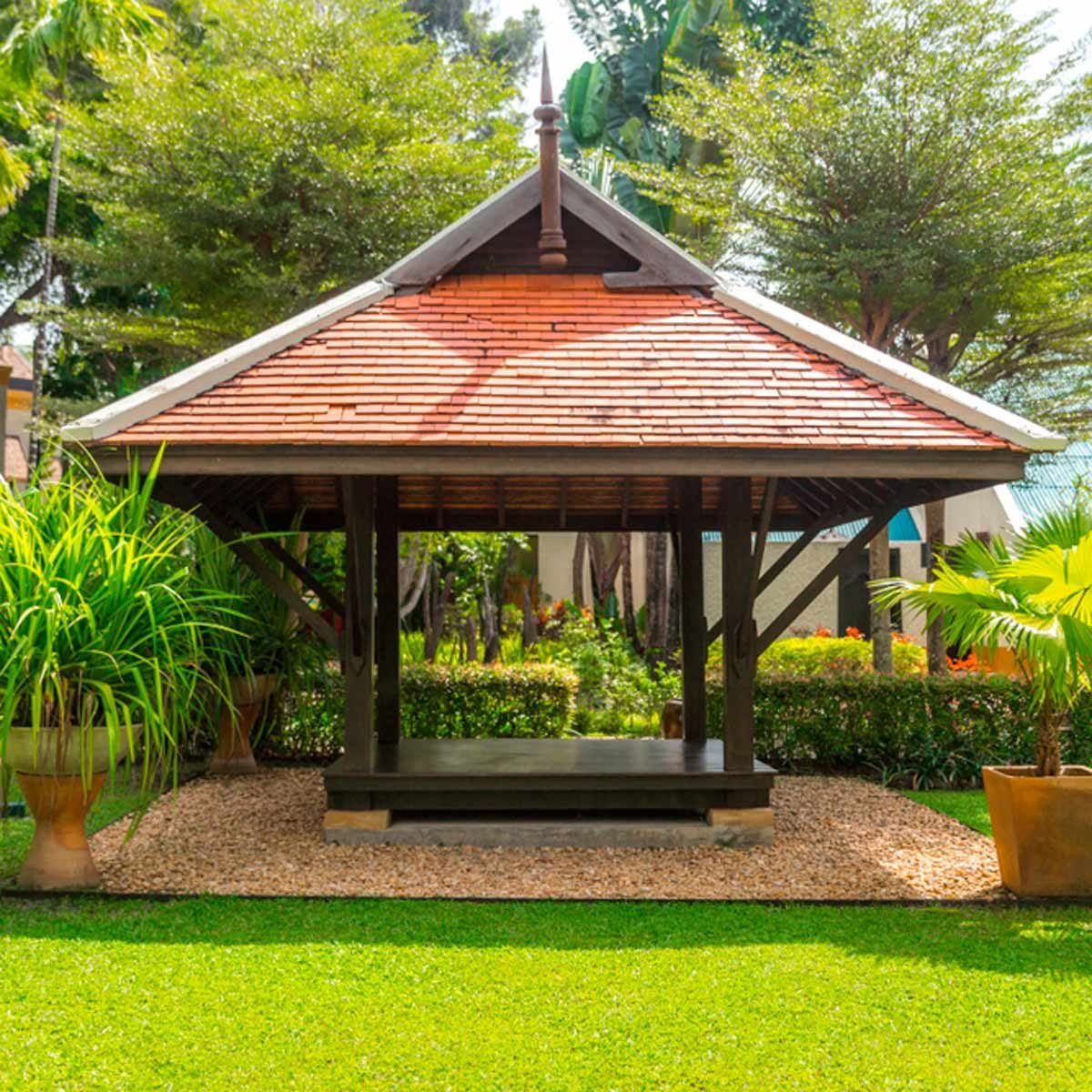 A wooden pavilion with a tiled roof sits on pebbled ground, surrounded by lush green grass and vibrant plants in a sunny garden setting.
