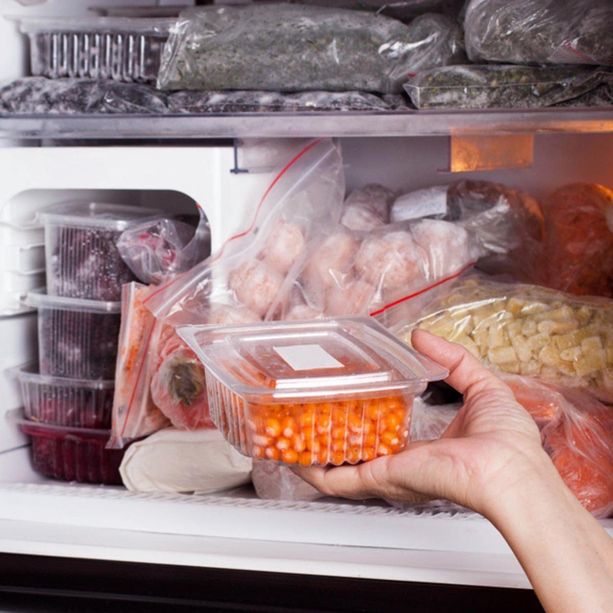 A hand holds a clear container of orange beads while reaching into a crowded freezer filled with various frozen foods in bags and containers