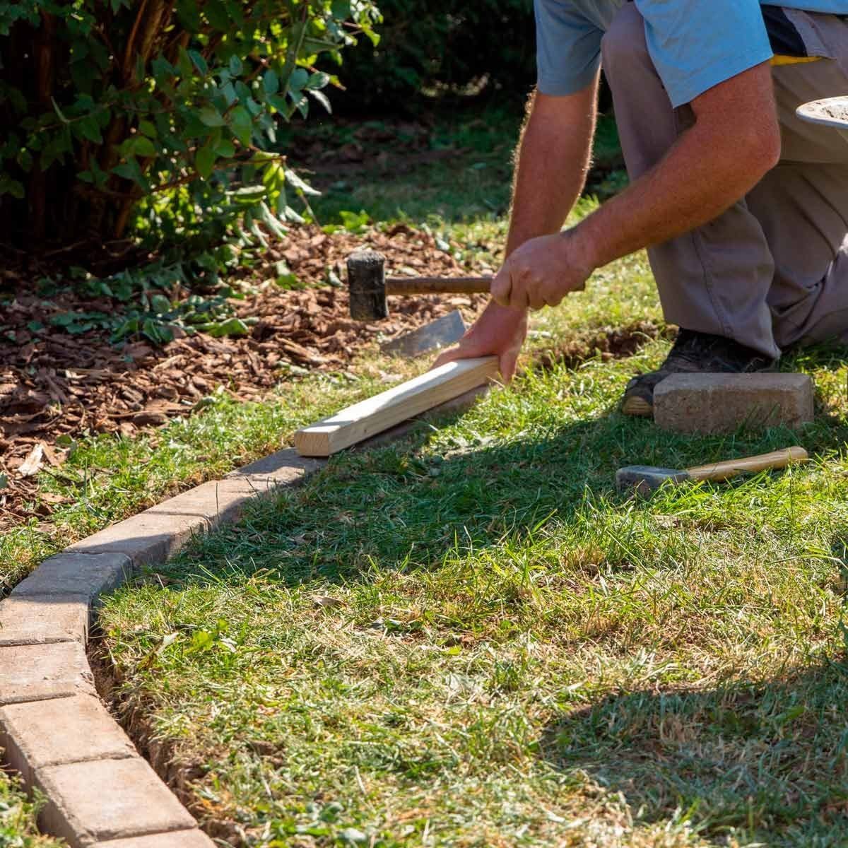 A person using a wooden tool strikes a brick pathway while working in a green garden, surrounded by plants and mulch on the ground.