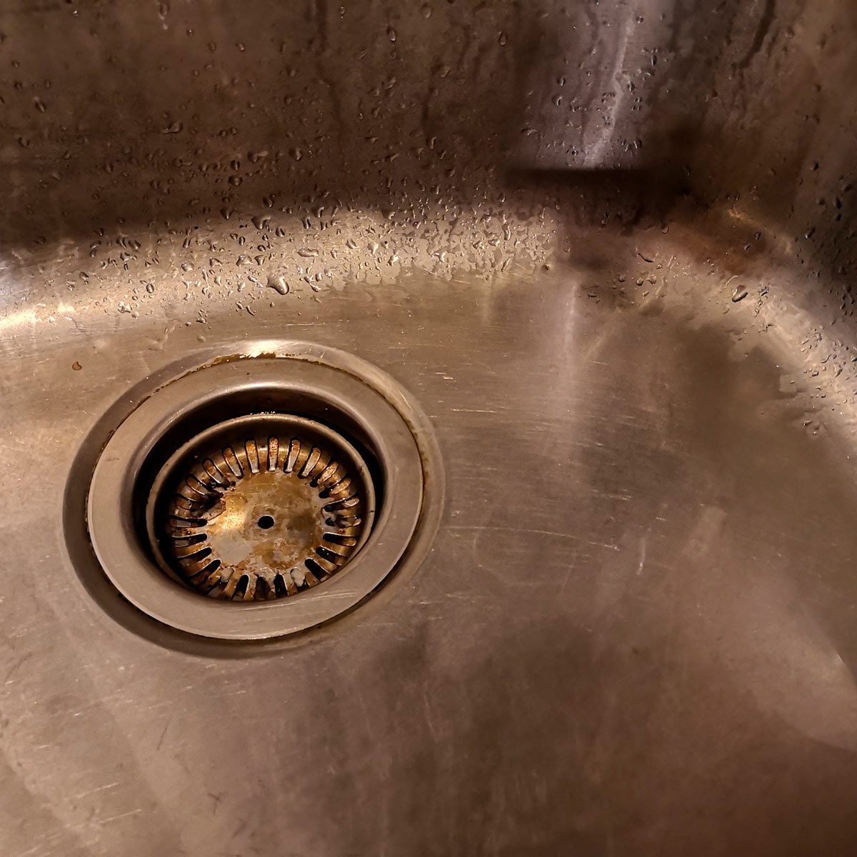 A metal sink contains a round drain with a grid, surrounded by droplets of water, reflecting light in a dimly lit environment.