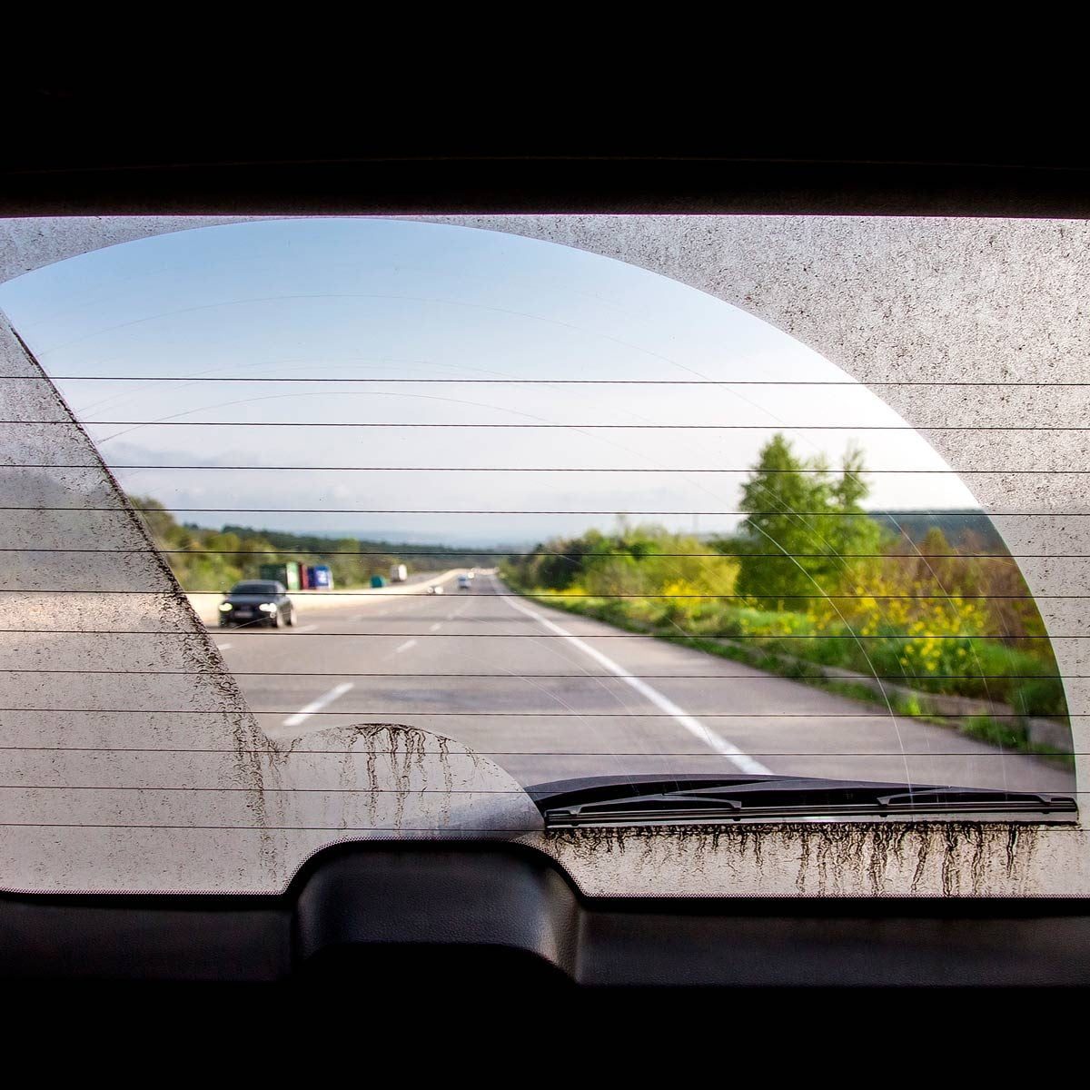 A rear view of a highway shows two cars traveling; the scene is partially obscured by a dirty back window with visible streaks.