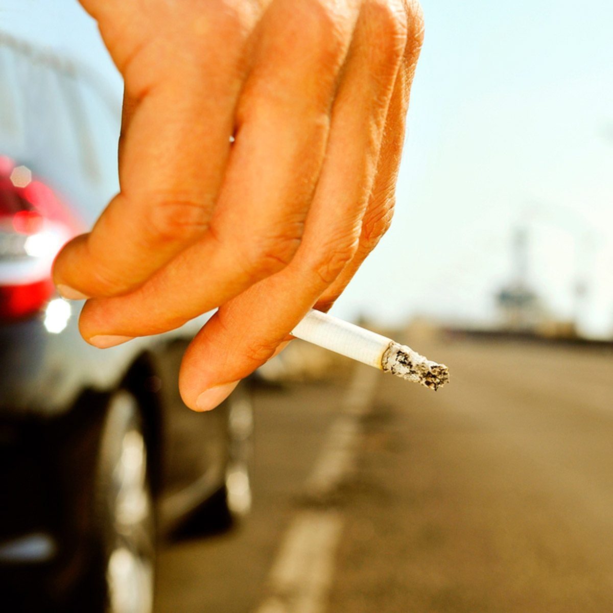 A hand holds a cigarette, releasing smoke while resting beside a parked car on a deserted road under a clear, bright sky.