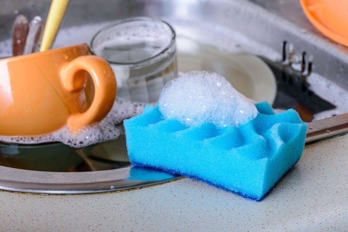 A blue sponge with soap bubbles rests on a counter near a orange cup and a glass, surrounded by soapy water in a sink.