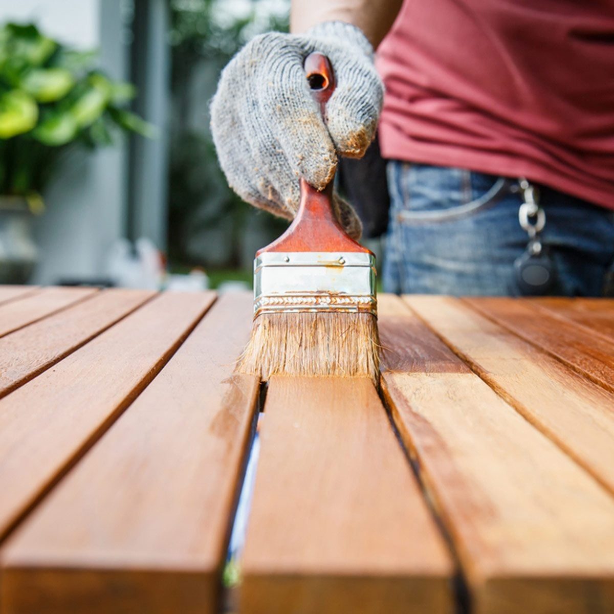 A gloved hand applies varnish with a brush to wooden slats on a table, surrounded by greenery in an outdoor setting.