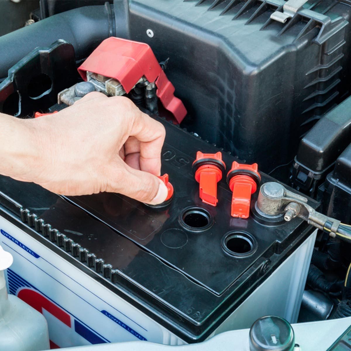 A hand removes a red cap from a car battery, surrounded by components of a vehicle engine.