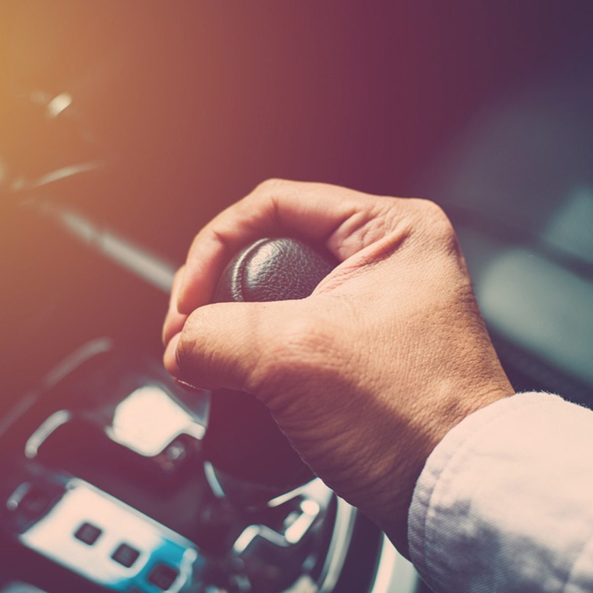 A hand grips a gear shift, preparing to change gears. The environment suggests a vehicle's interior, with subtle light creating a warm atmos