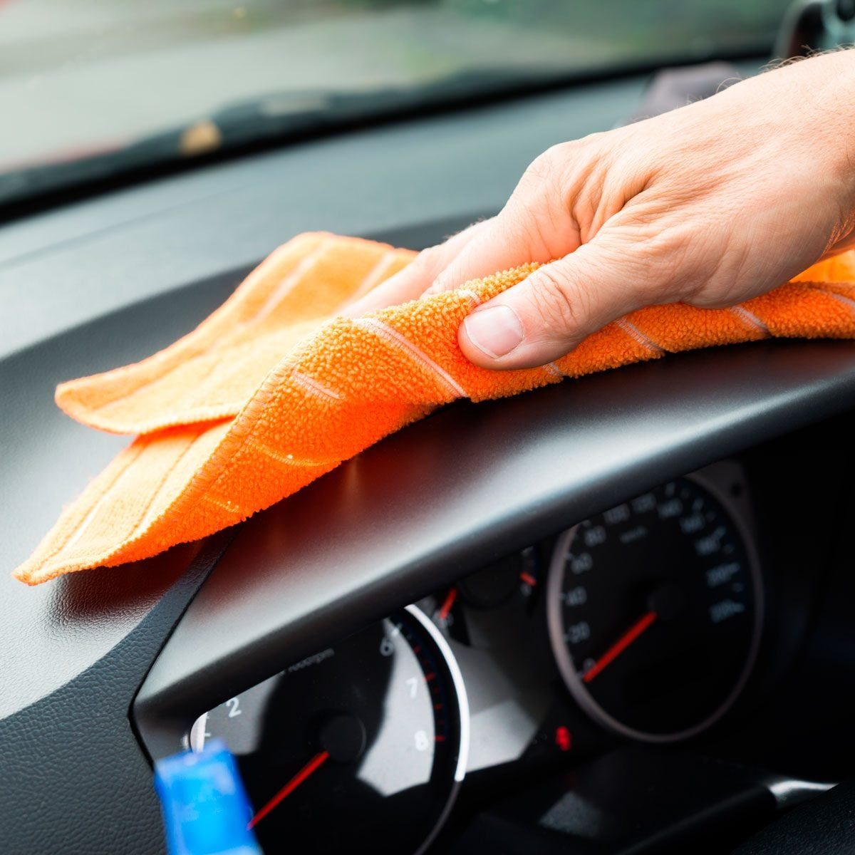 A hand wipes a dark dashboard with an orange cleaning cloth inside a car, surrounded by dashboard gauges and bright lighting.