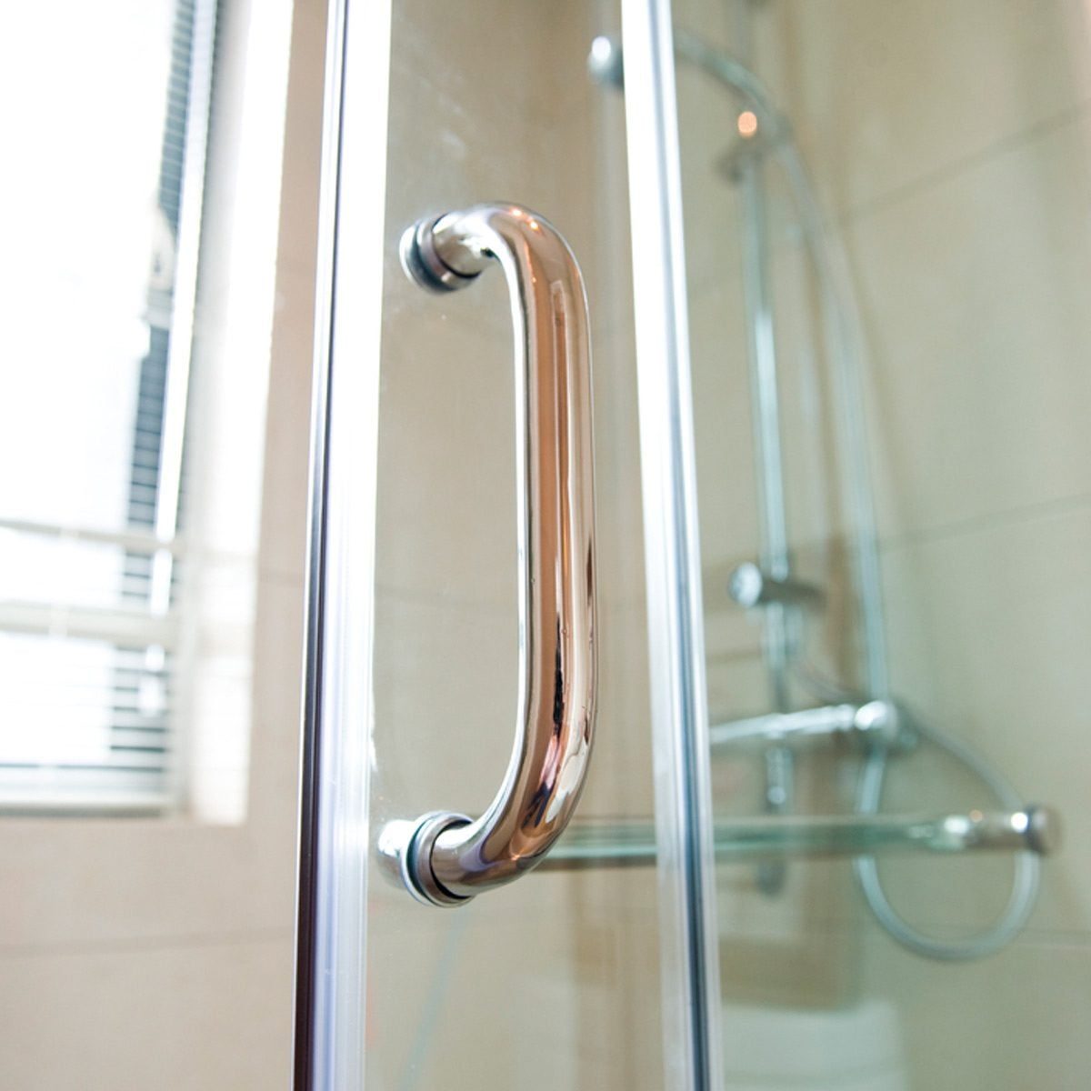 A polished metal handle is mounted on a glass shower door, providing grip. Soft light filters through a nearby window, illuminating tiled wa