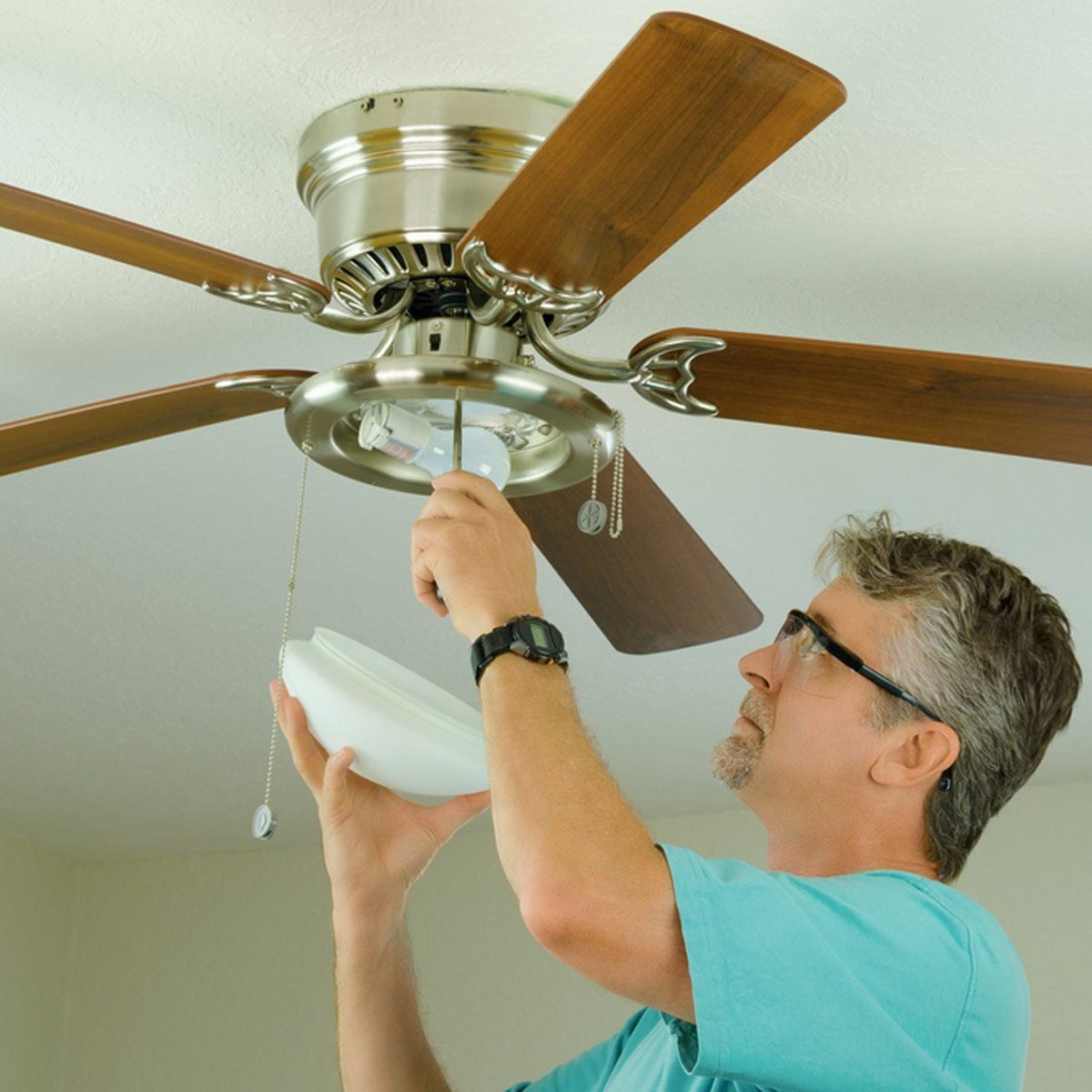 A man is installing or repairing a ceiling fan while holding a light cover, indoors, with wooden blades and a metallic base.