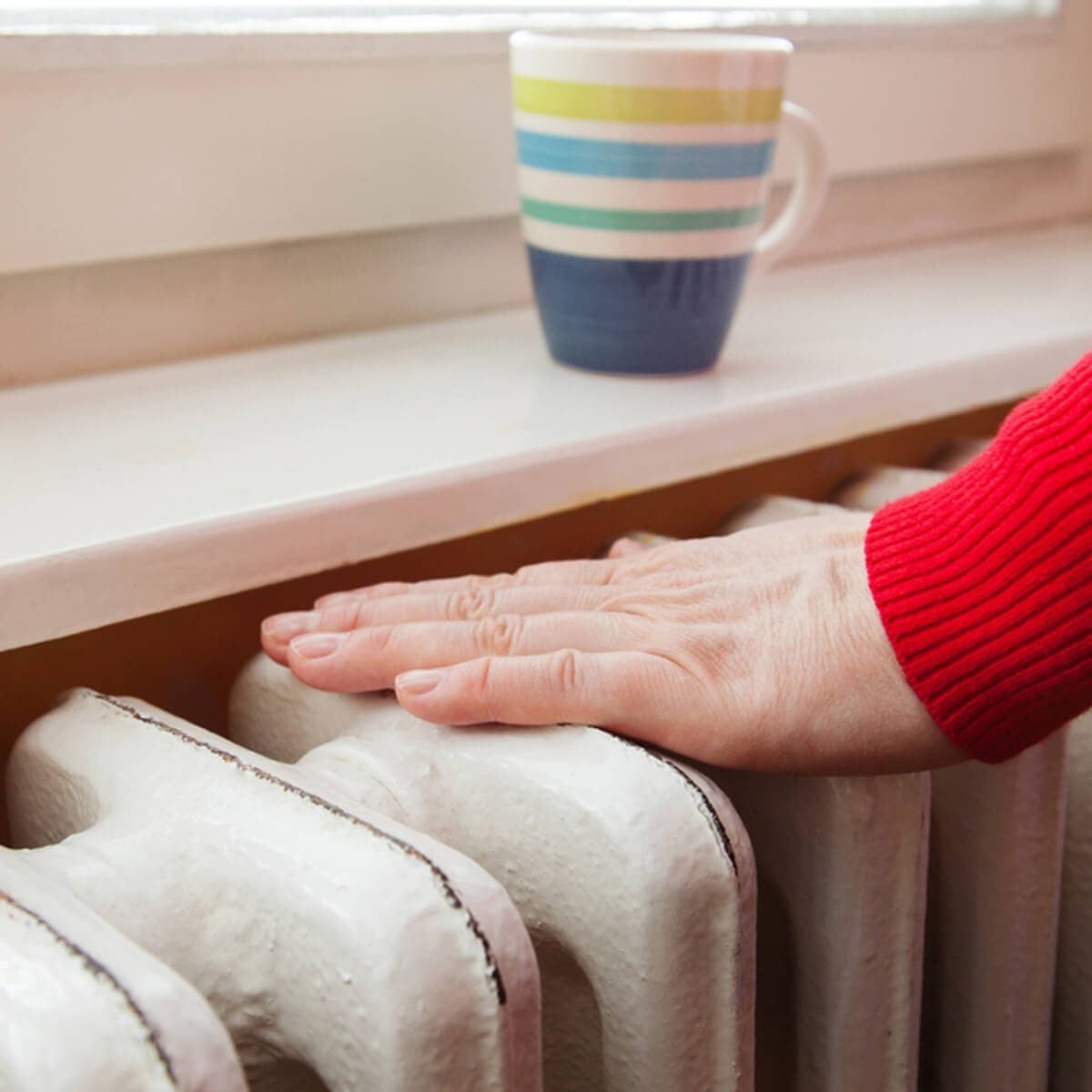 A hand rests on a white radiator, while a striped mug sits nearby on a windowsill, suggesting a cozy indoor setting.
