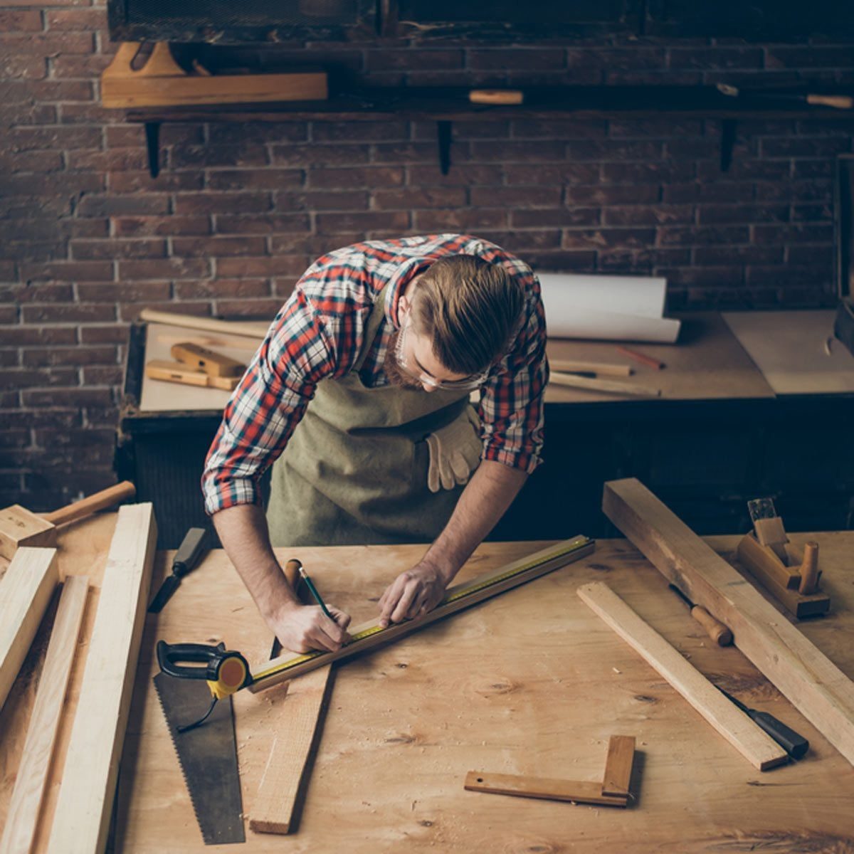 A man measures and marks wood on a workbench in a rustic workshop, surrounded by various tools and lumber on a wooden surface.