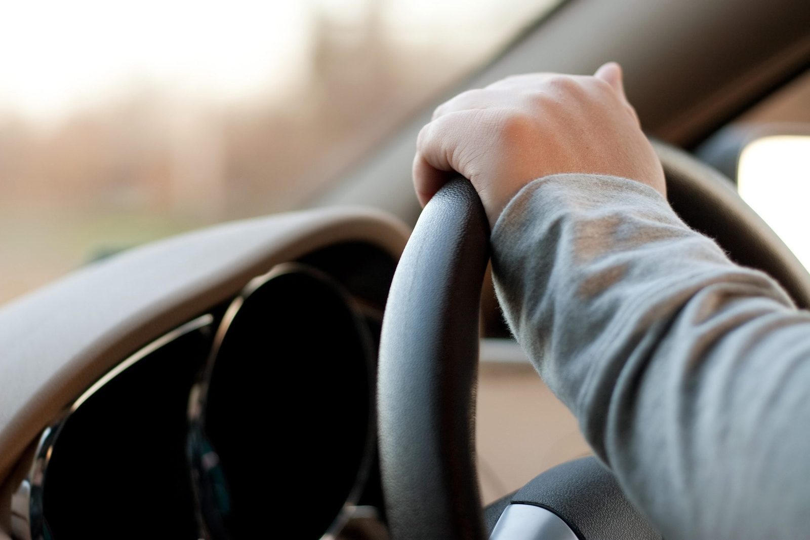 A hand grips a steering wheel, maneuvering a vehicle inside a car, with blurred surroundings suggesting movement and an outdoor setting.