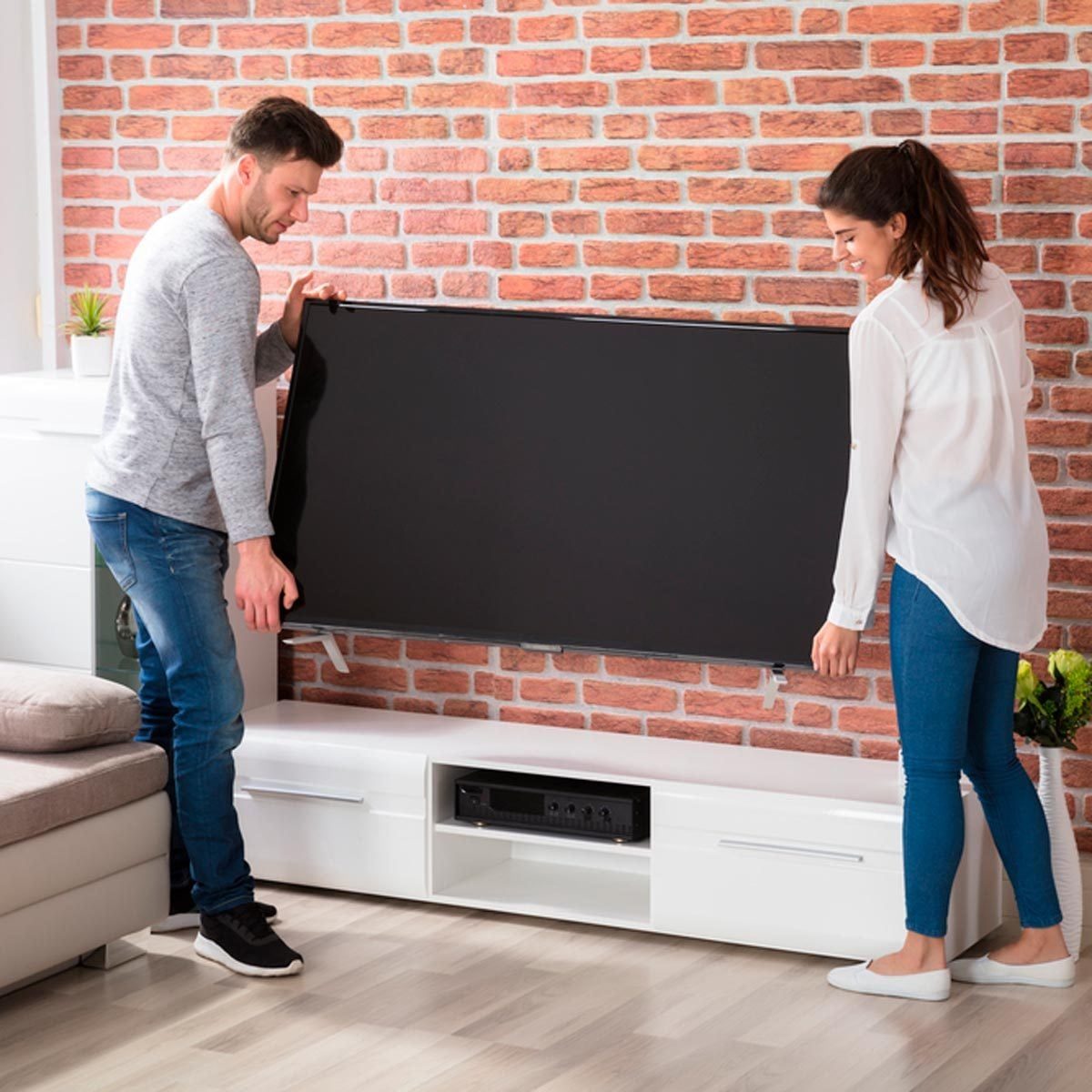 Two people hold a black television, preparing to set it up against a brick-patterned wall in a modern, brightly lit room.