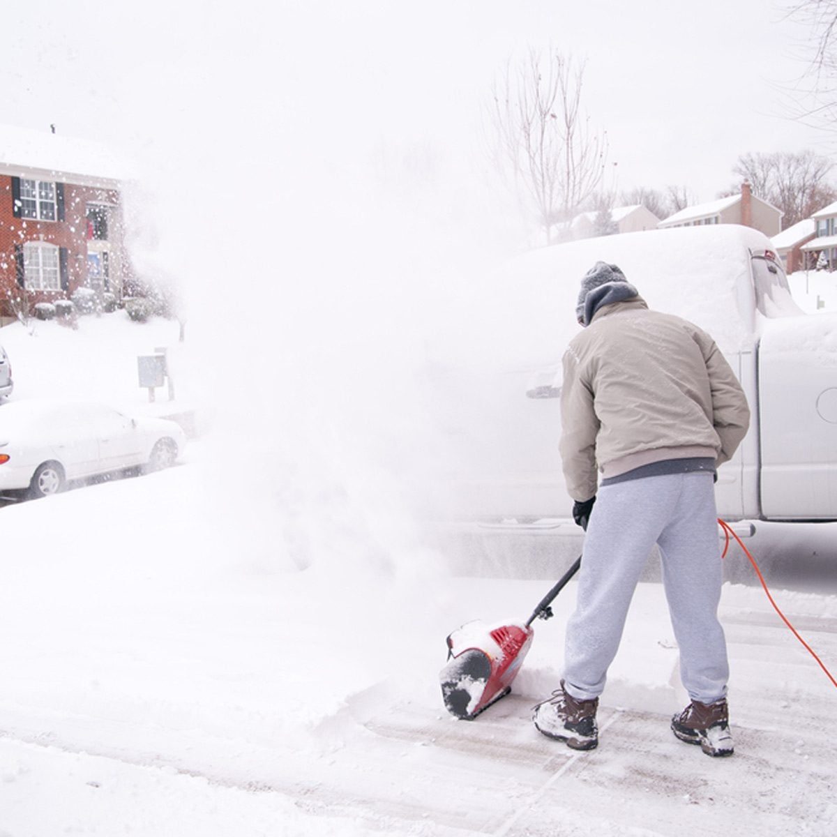 A person operates a snow blower, clearing a driveway covered in fresh snow, while residential houses and a car are visible in the snowy back