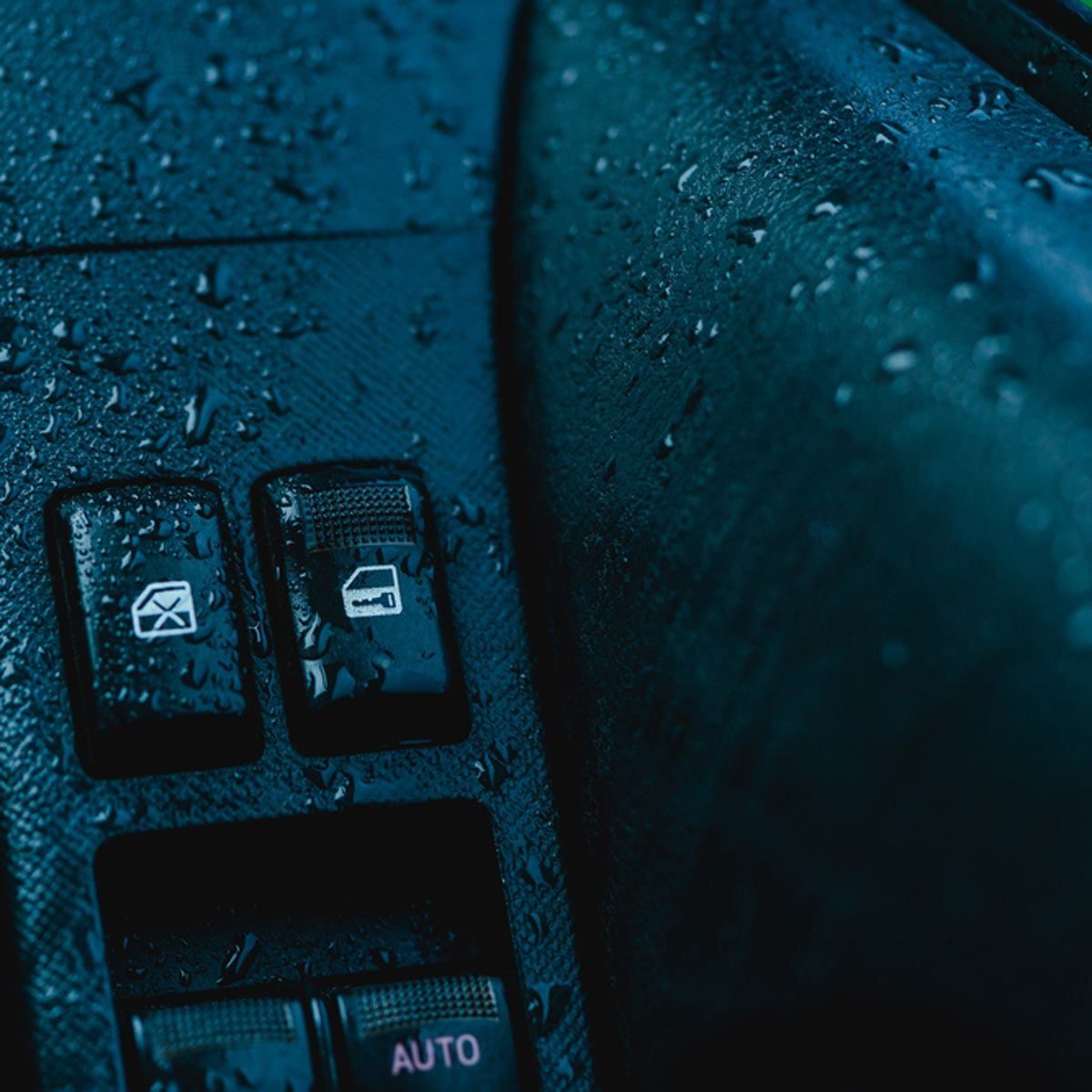 Rainy droplets rest on a car door panel, focusing on two control buttons for windows or locks, suggesting a damp interior environment.