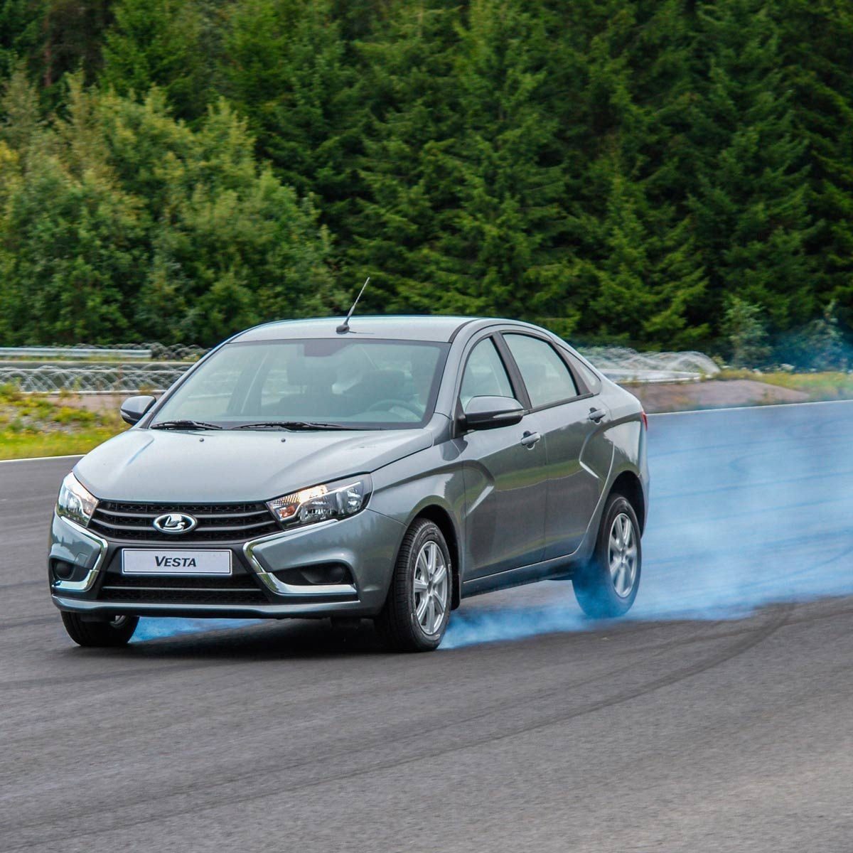 A gray sedan skids on a track, emitting blue smoke, surrounded by a green forest backdrop. The car is labeled "Vesta."
