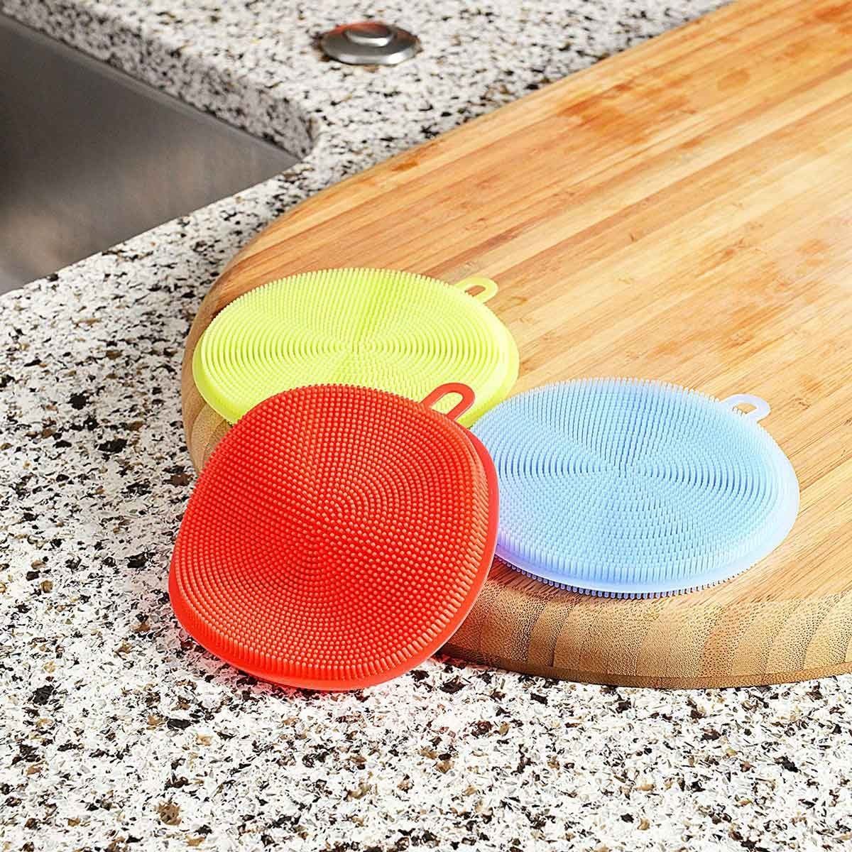 Colorful silicone dish scrubbers rest on a wooden cutting board beside a sink, against a speckled countertop, showcasing various textures an