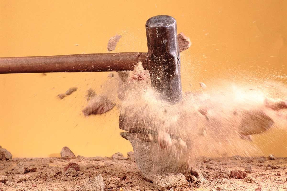 A hammer strikes a rock, creating dust and debris that disperse in the air, set against a smooth, yellow background.