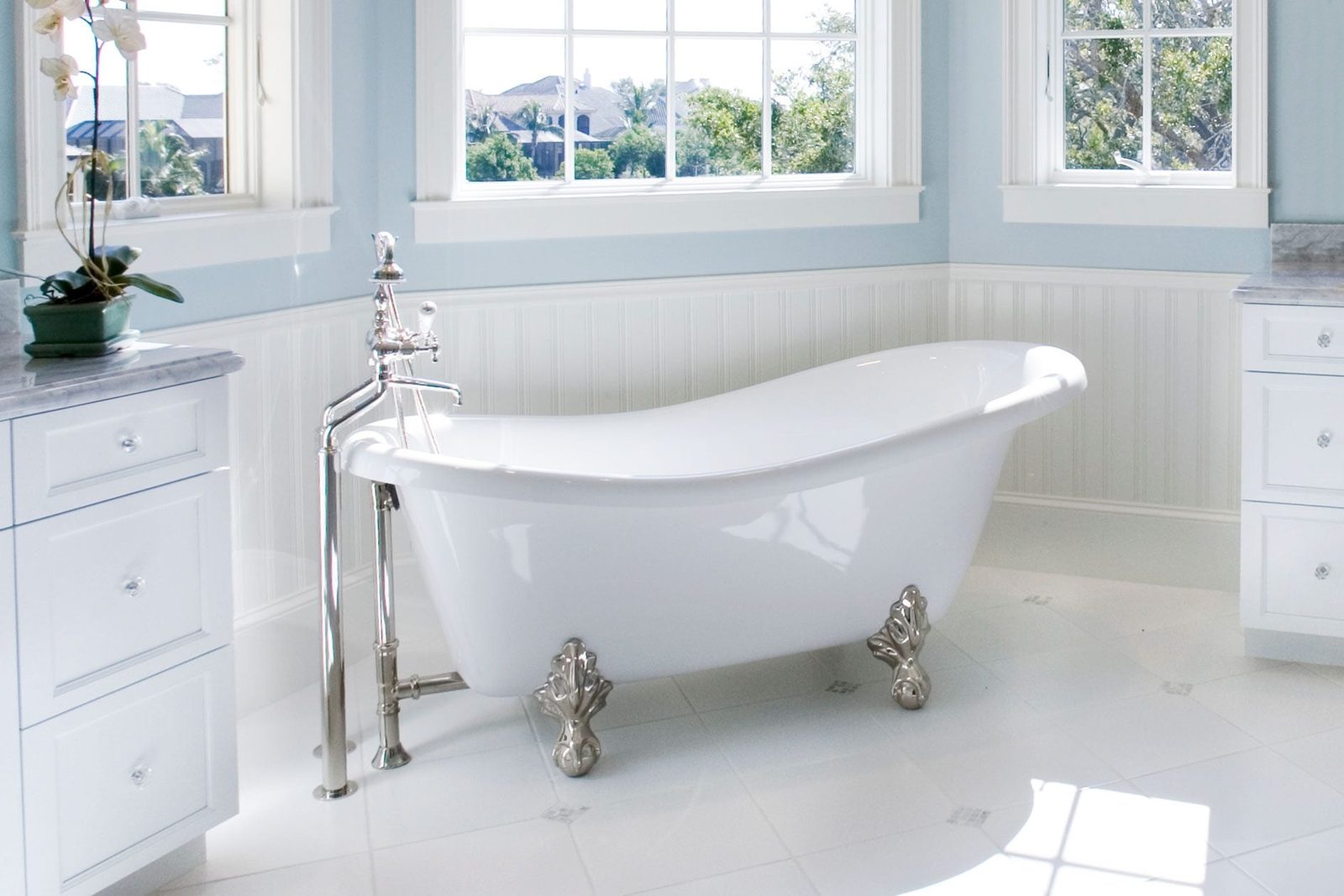 A freestanding white bathtub rests on ornate silver feet, surrounded by light blue walls and natural light from large windows, creating a se