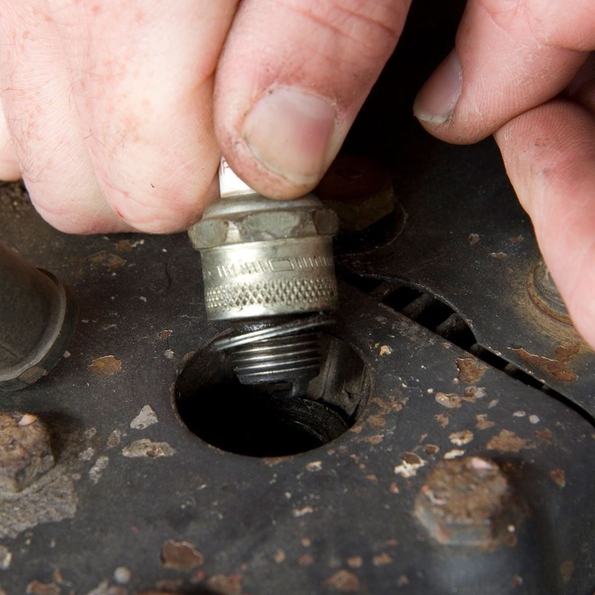 A hand grips a spark plug, twisting it into an engine block's threaded opening, amidst a dirty, mechanical environment with rusted bolts and