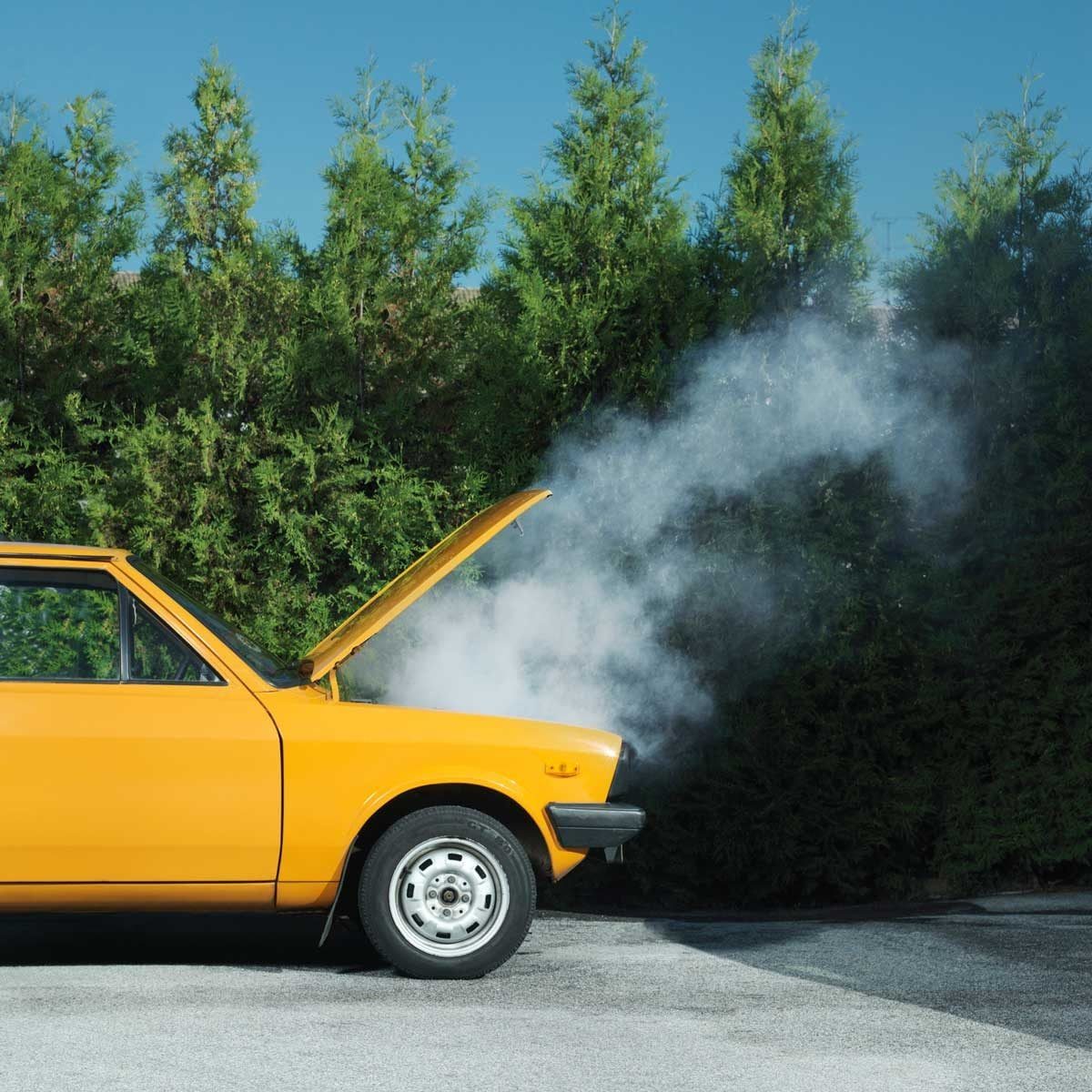 A yellow car with its hood raised is emitting smoke, parked beside a dense green hedge under a clear blue sky.