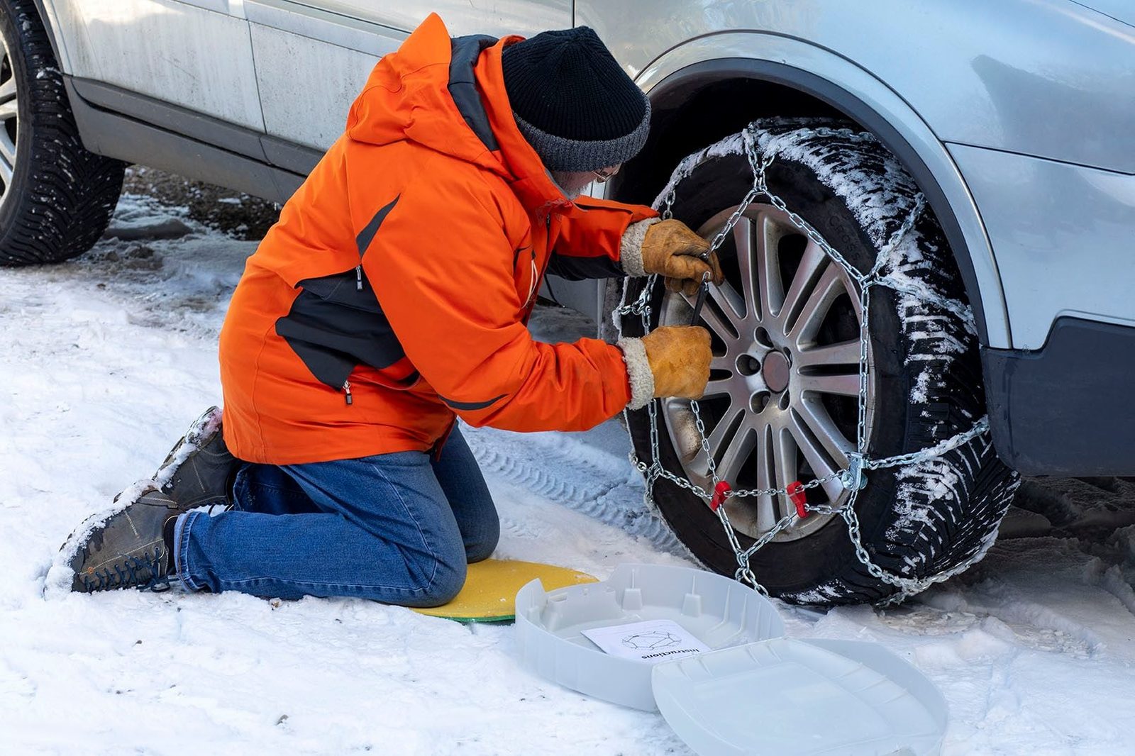 A person in an orange jacket attaches snow chains to a car tire while kneeling on snow-covered ground. A white container sits nearby.