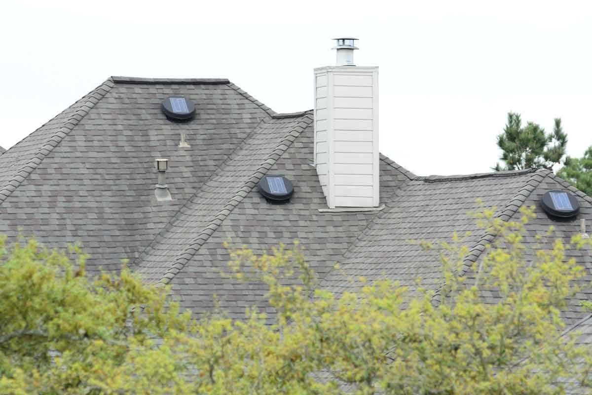 A sloped roof features multiple solar panels and a chimney, surrounded by green foliage and a cloudy sky in the background.
