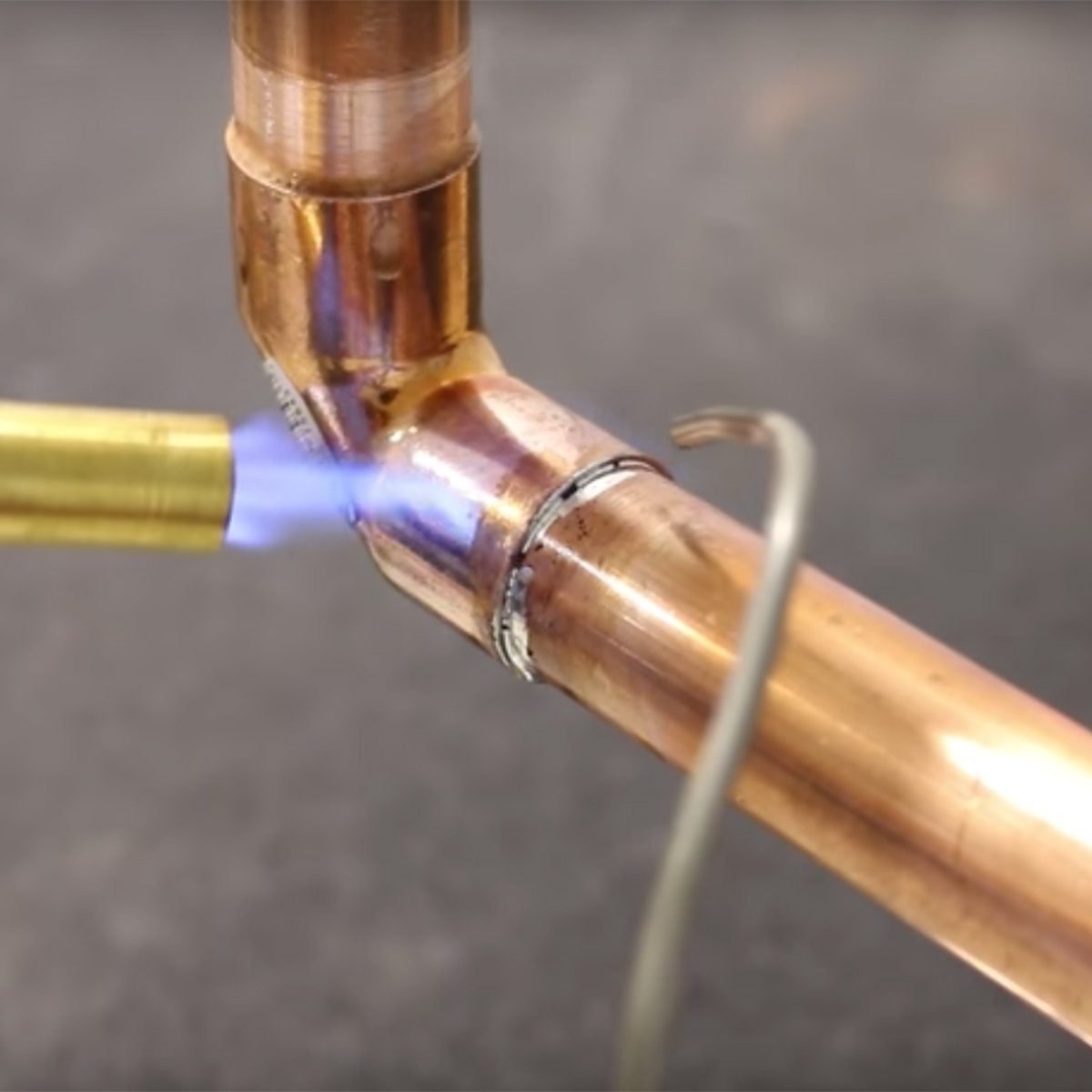 A gas torch emits a blue flame, melting solder at a joint where two copper pipes connect, in a workshop environment.