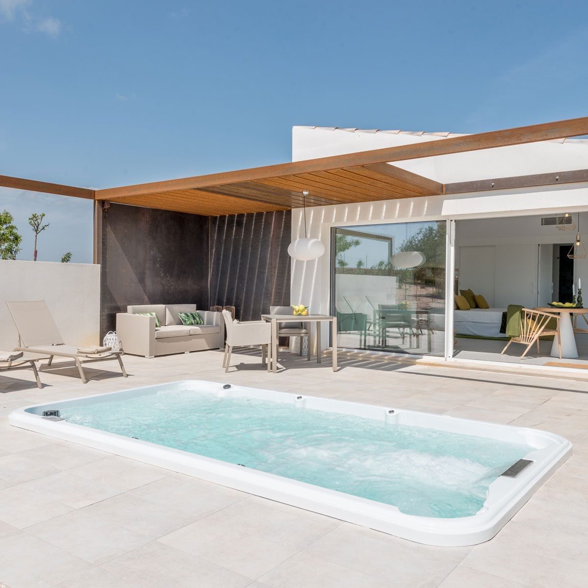 A rectangular hot tub bubbles in a sunlit patio. Nearby, a sofa and dining table set the scene for a relaxed outdoor gathering.