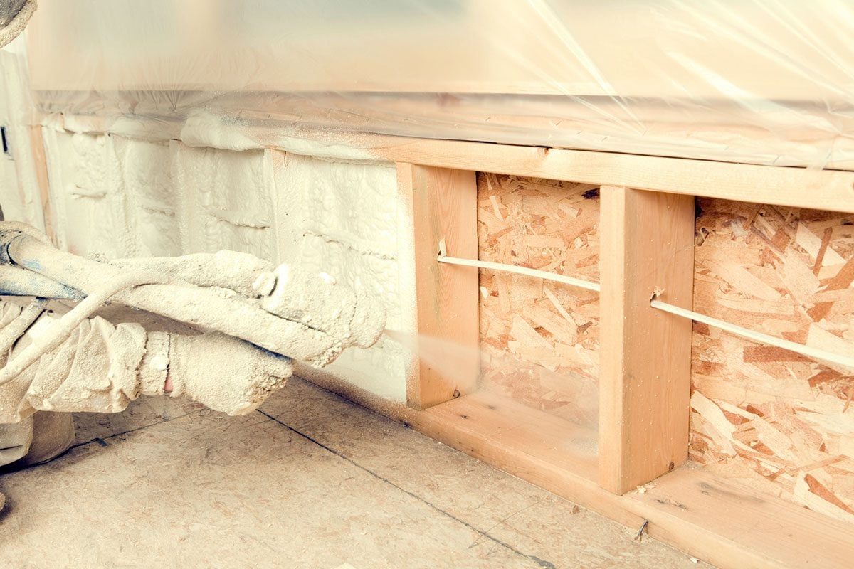 A person applies spray foam insulation to wall cavities in a construction area, surrounded by wooden frames and plastic sheeting to contain