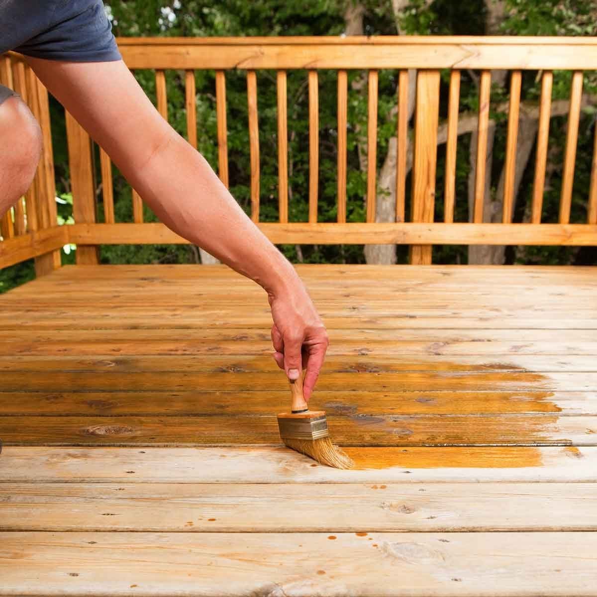 A person is applying a wood stain with a brush onto a wooden deck, surrounded by greenery and wooden railings.