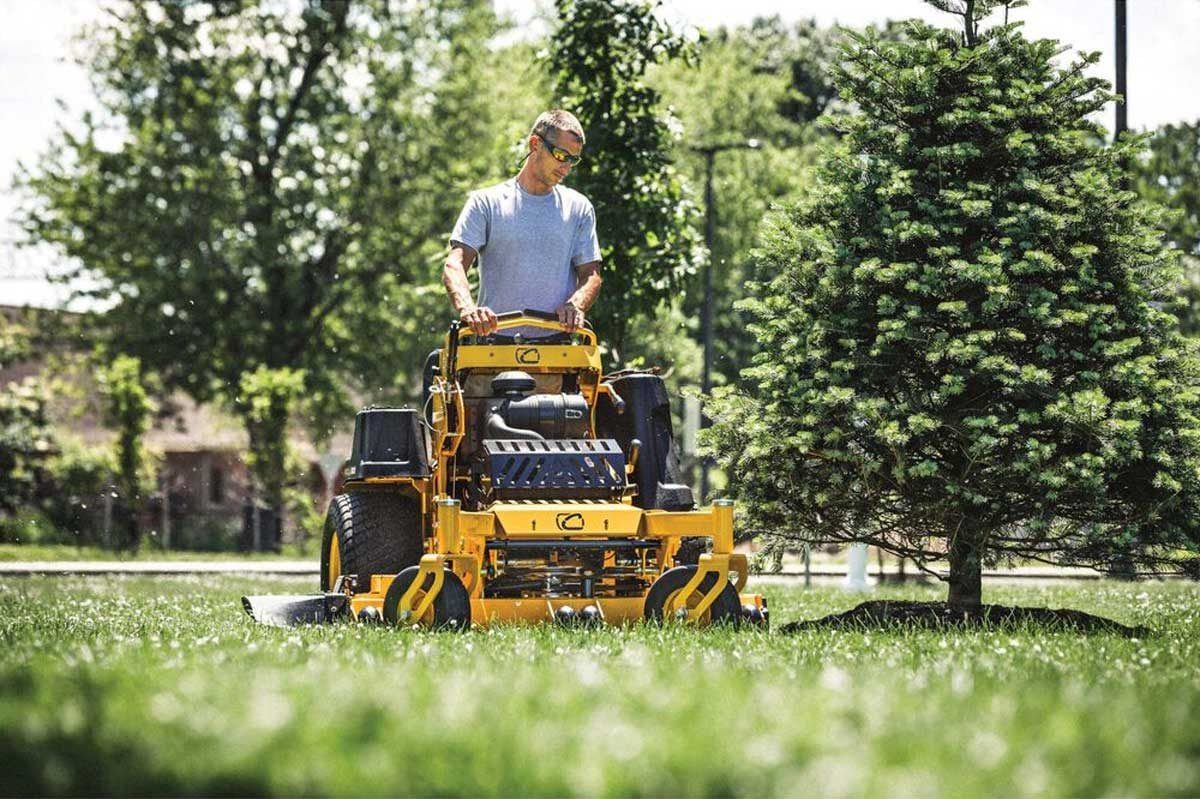A man operates a yellow zero-turn mower, cutting grass near a tree in a sunny park with lush greenery surrounding him.