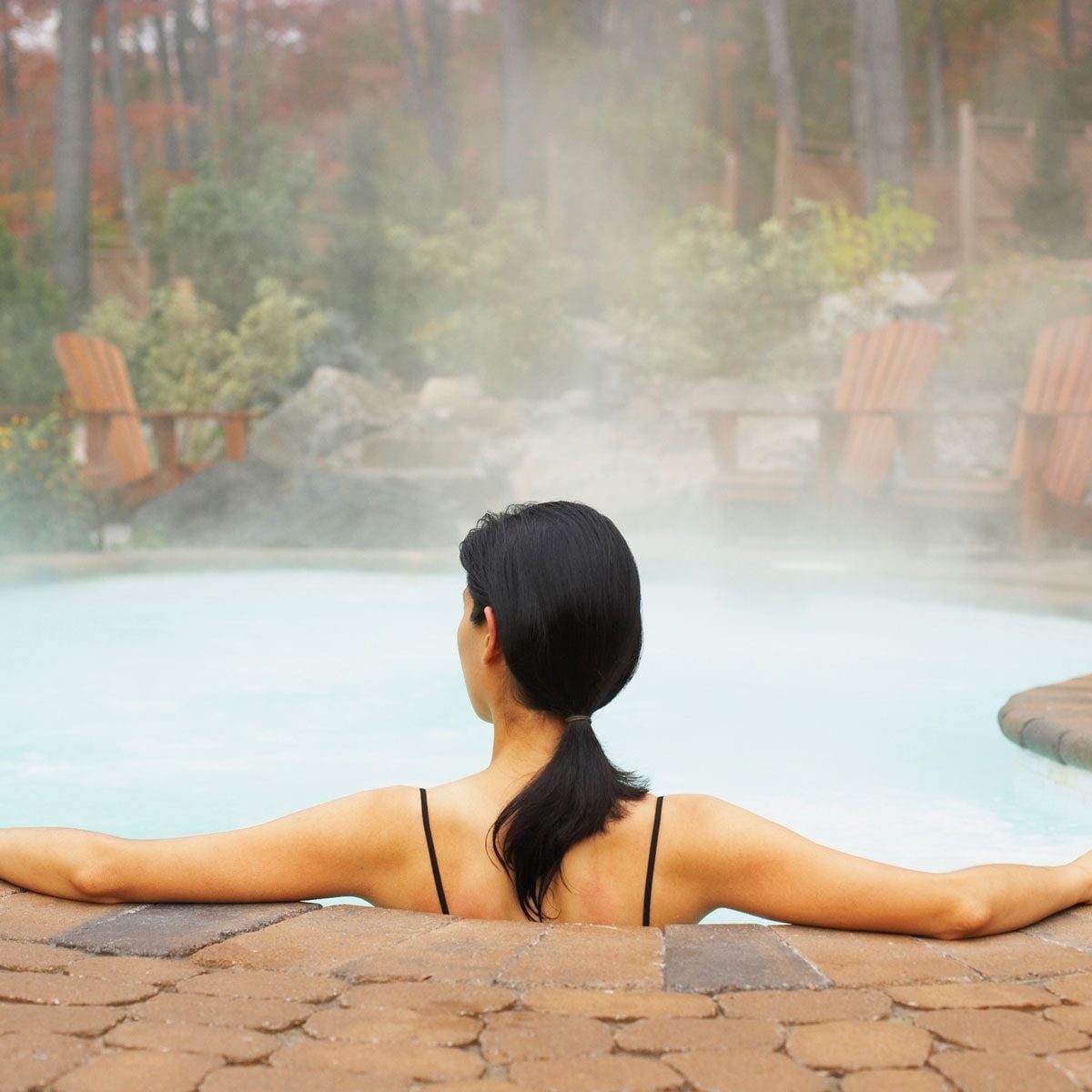 A woman relaxes in a steaming hot tub, her arms outstretched, surrounded by mist and greenery, with wooden chairs in the background.