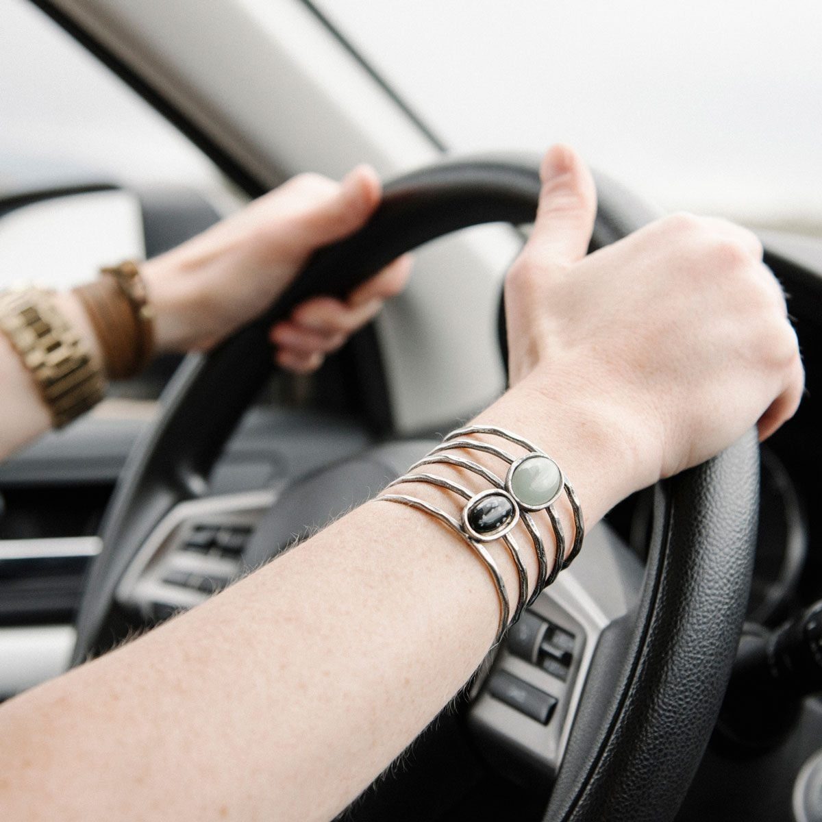 A hand grips a steering wheel, adorned with a decorative bracelet, while driving in a vehicle, indicating motion and active engagement on th
