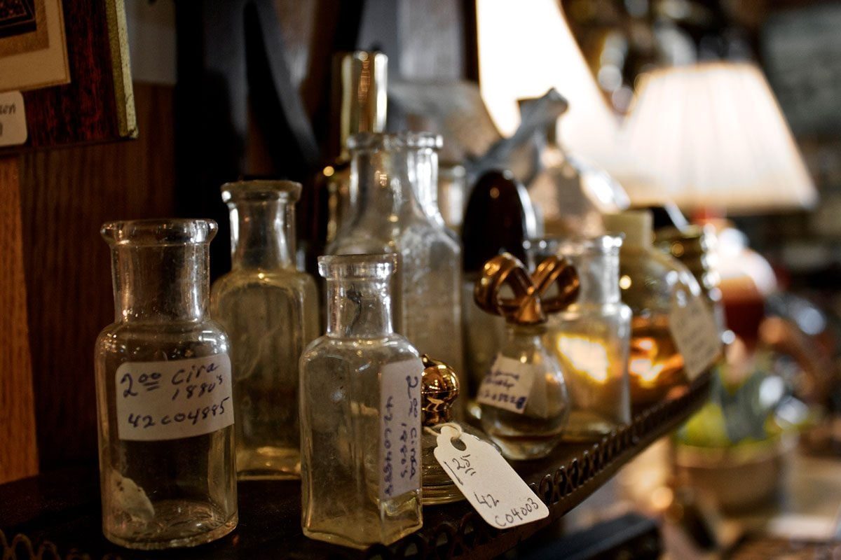 Glass bottles are arranged on a wooden shelf, some with price tags, while a warm ambient light illuminates the surrounding vintage decor.