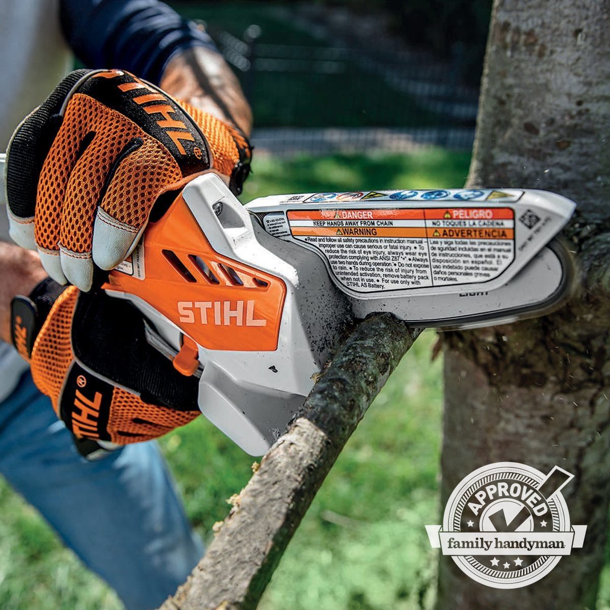 A person using a battery-powered chainsaw to cut a tree branch, surrounded by grass and trees, wearing protective gloves.