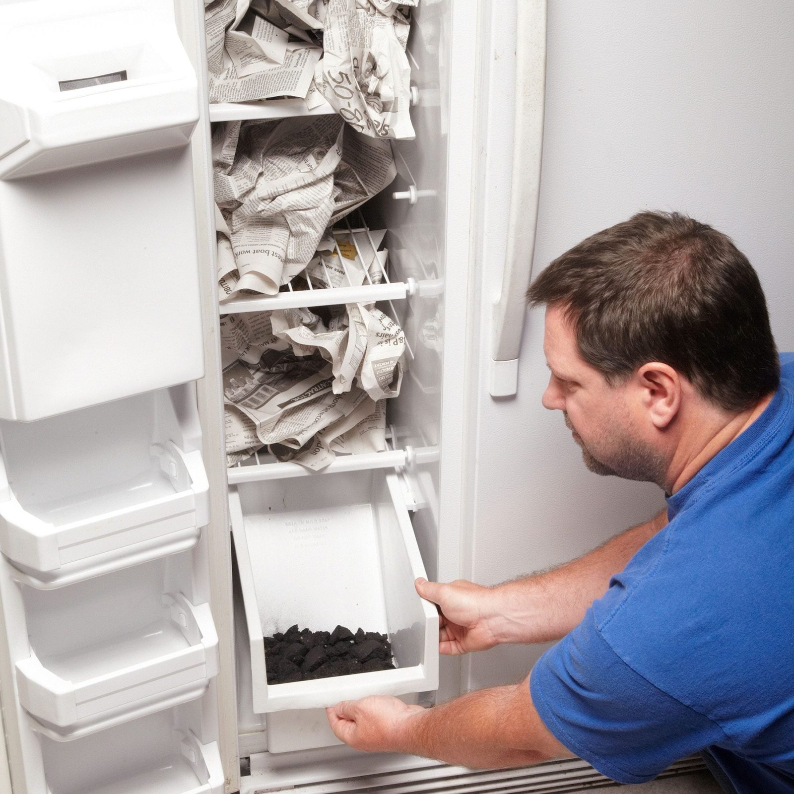 A man pulls out a drawer filled with black material from a white refrigerator, while the interior displays crumpled newspaper among its shel