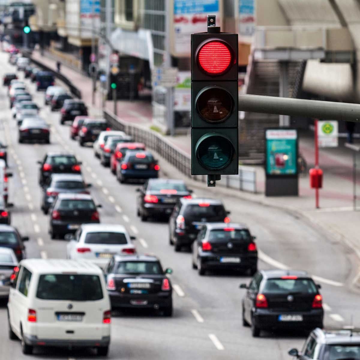 A red traffic light signals vehicles to stop on a busy road, where multiple cars are lined up, waiting in a city environment.