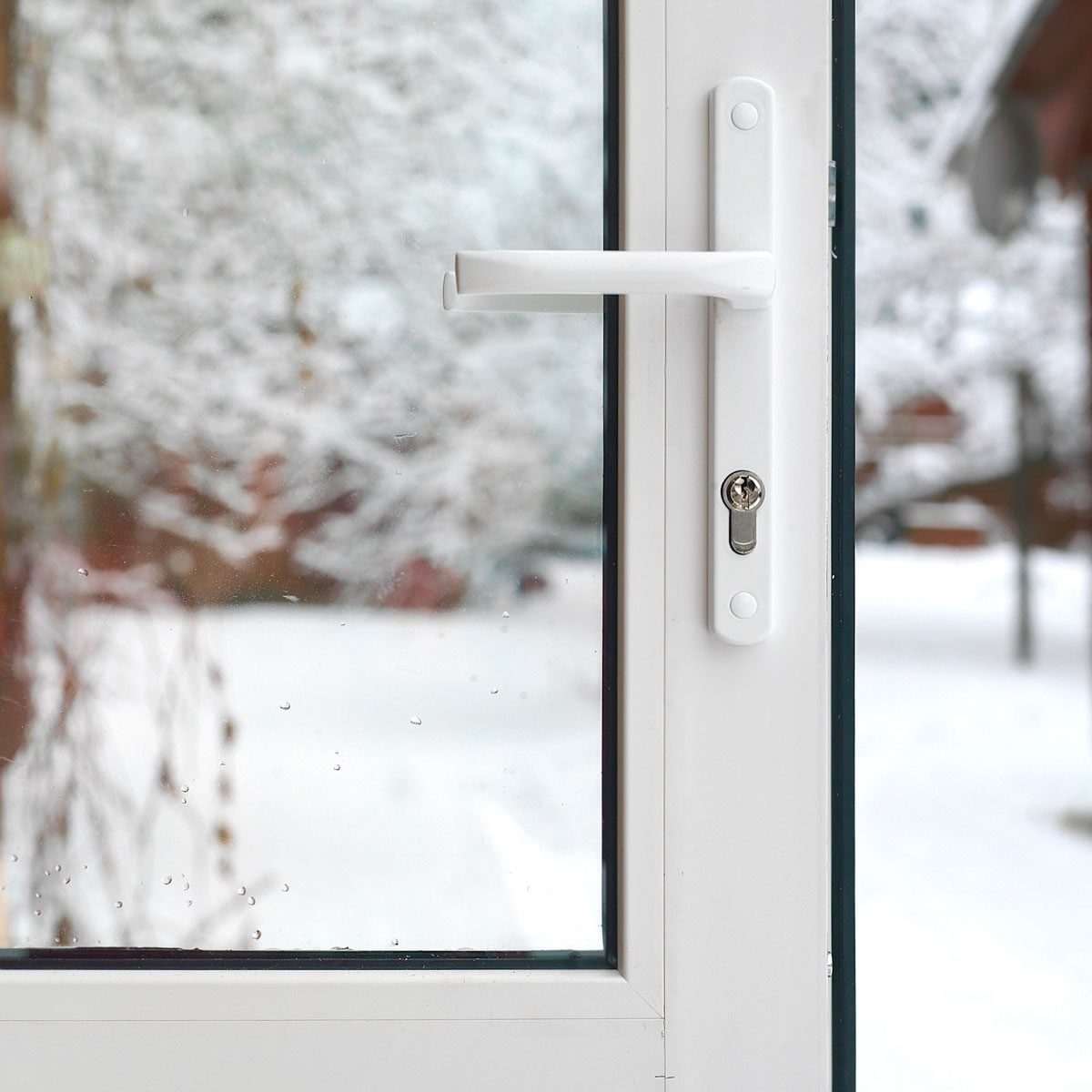 A white window handle is positioned by clear glass, showcasing a snowy landscape outside, with blurred trees and soft snowflakes visible in