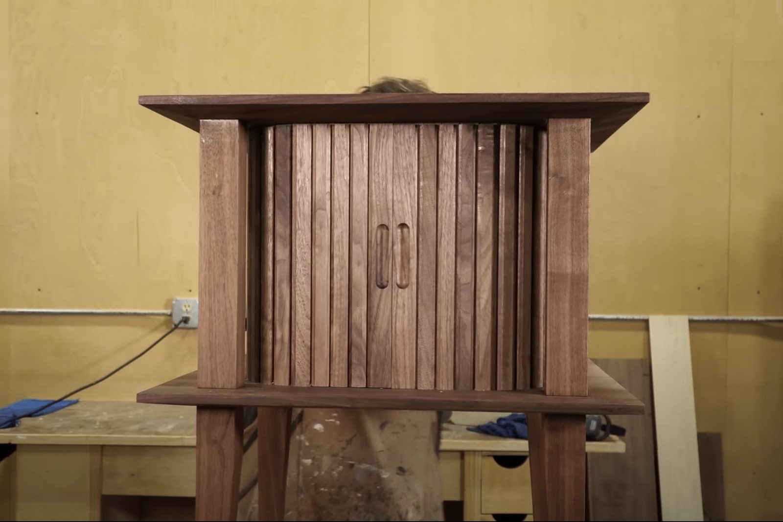 A wooden cabinet stands upright, showcasing vertical slats; a person partially obscured behind it is working in a workshop environment with