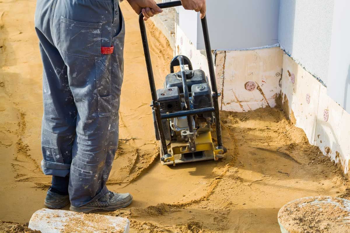 A worker operates a compactor on sand, preparing a construction site while standing on a flat surface near a building foundation.