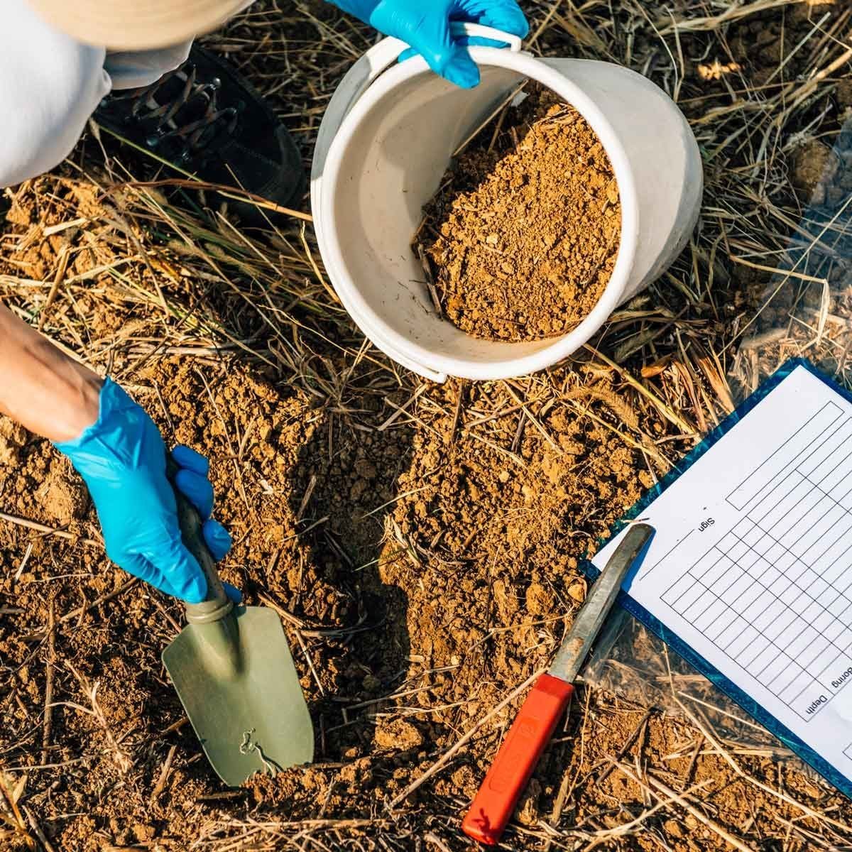A gloved hand uses a trowel to dig soil, placing it into a bucket on the ground, next to a clipboard with data sheets.