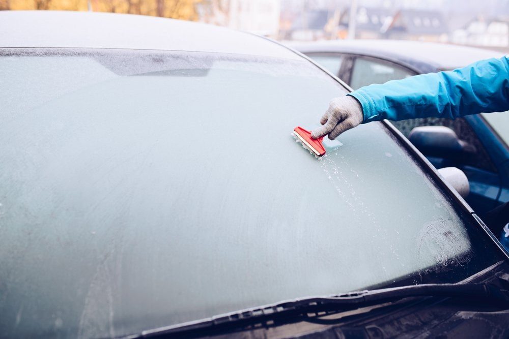 A person in a blue jacket uses a scraper to remove ice from a car windshield in a cold outdoor environment.