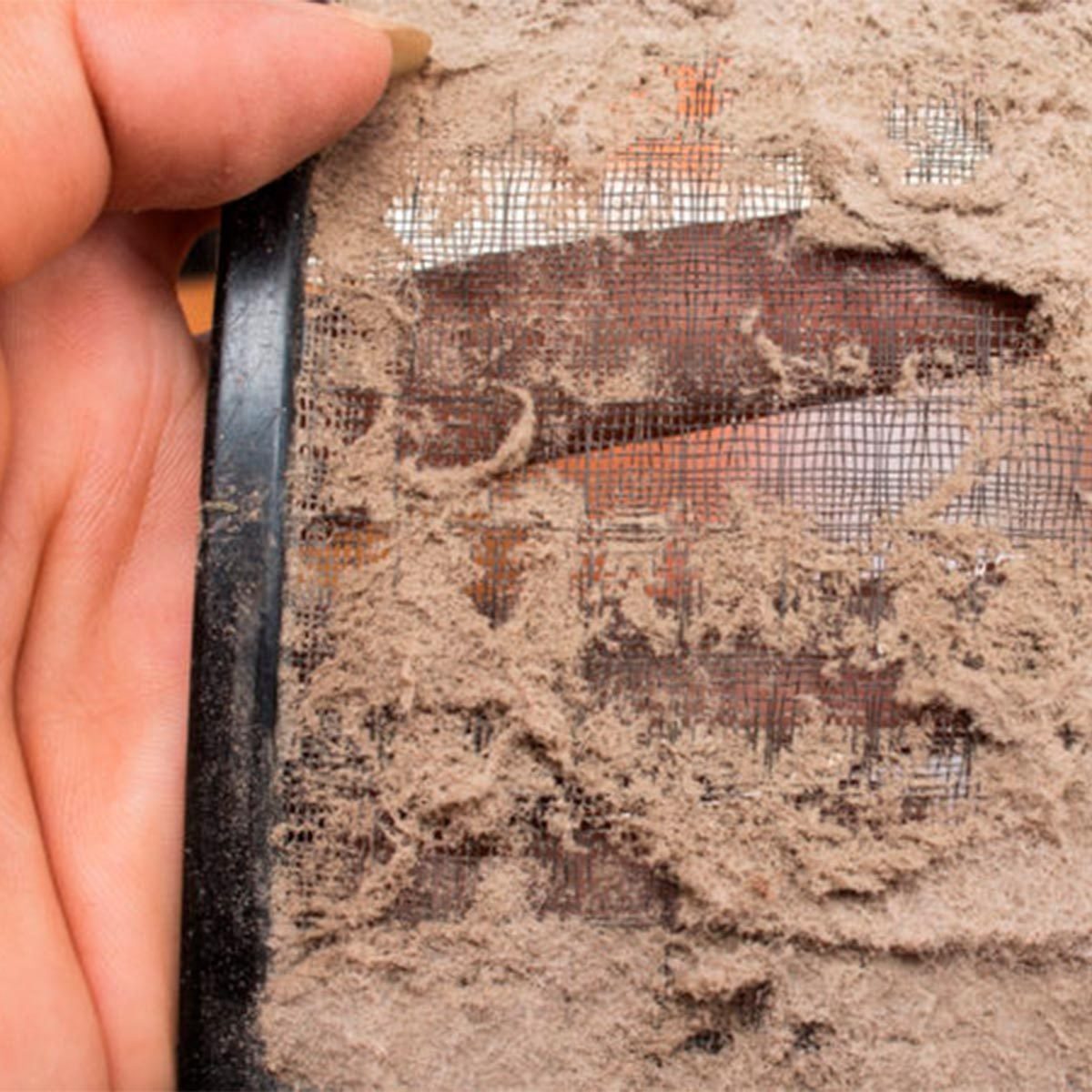 A hand holds a dusty air filter, showing a thick layer of dirt covering its mesh surface against a blurred background.
