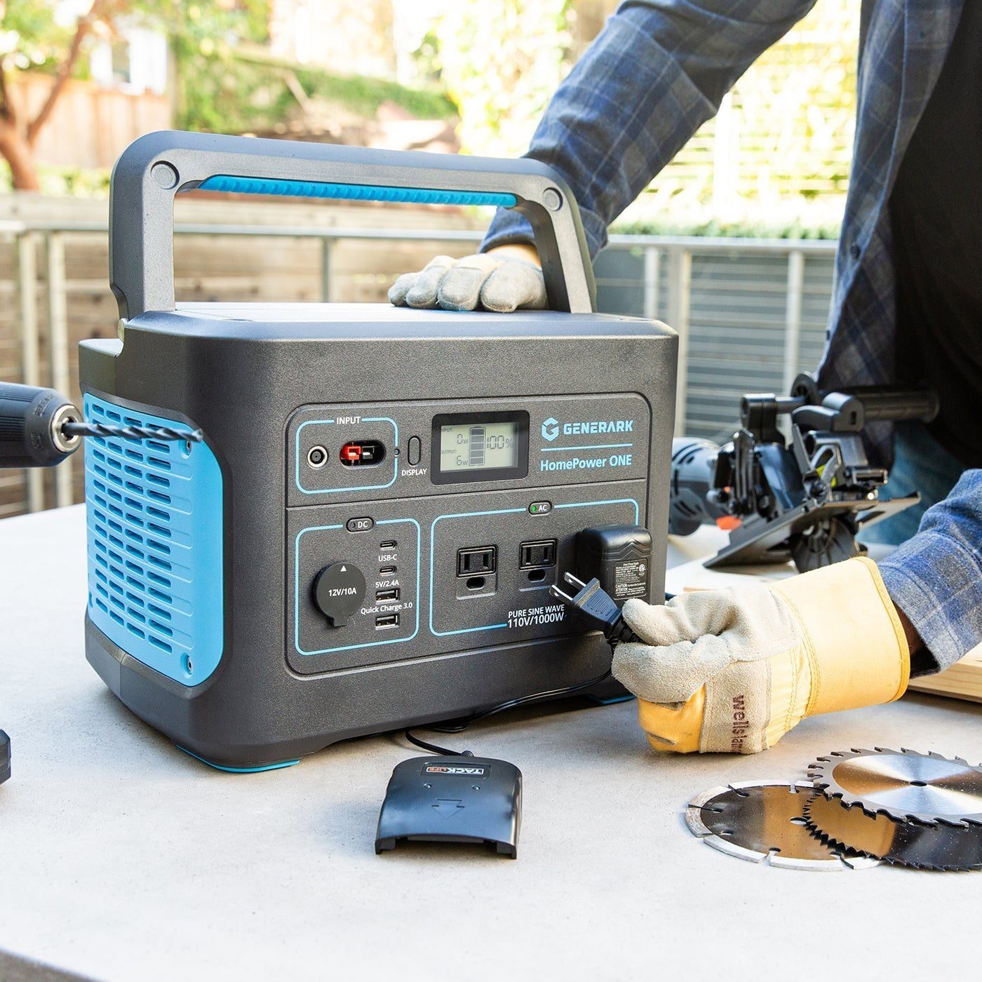 A portable power station is being connected to an electronic device on a worktable, surrounded by tools and saw blades in a backyard setting