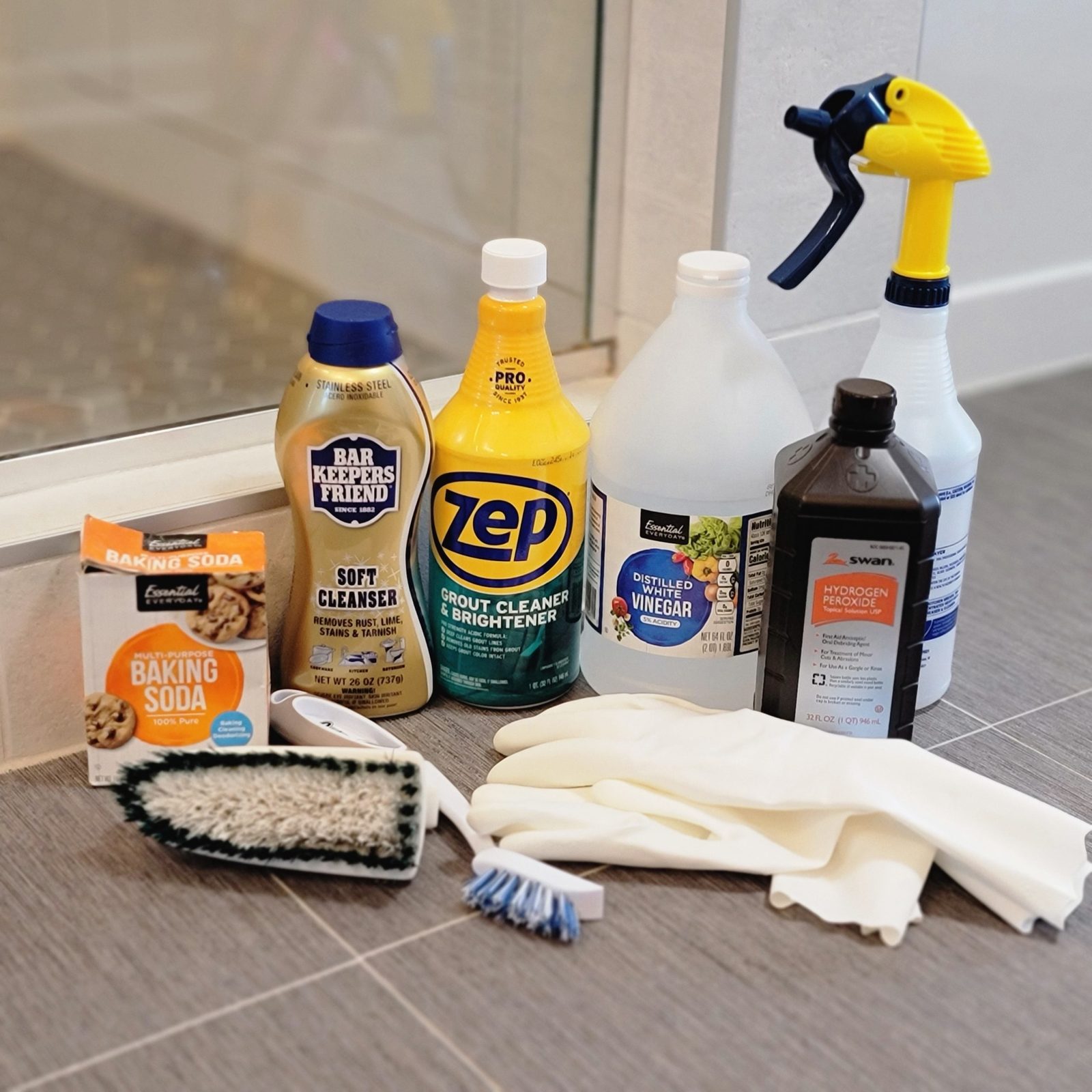 Cleaning supplies including various cleaners, baking soda, brushes, and gloves are arranged on a tiled floor near a glass shower.