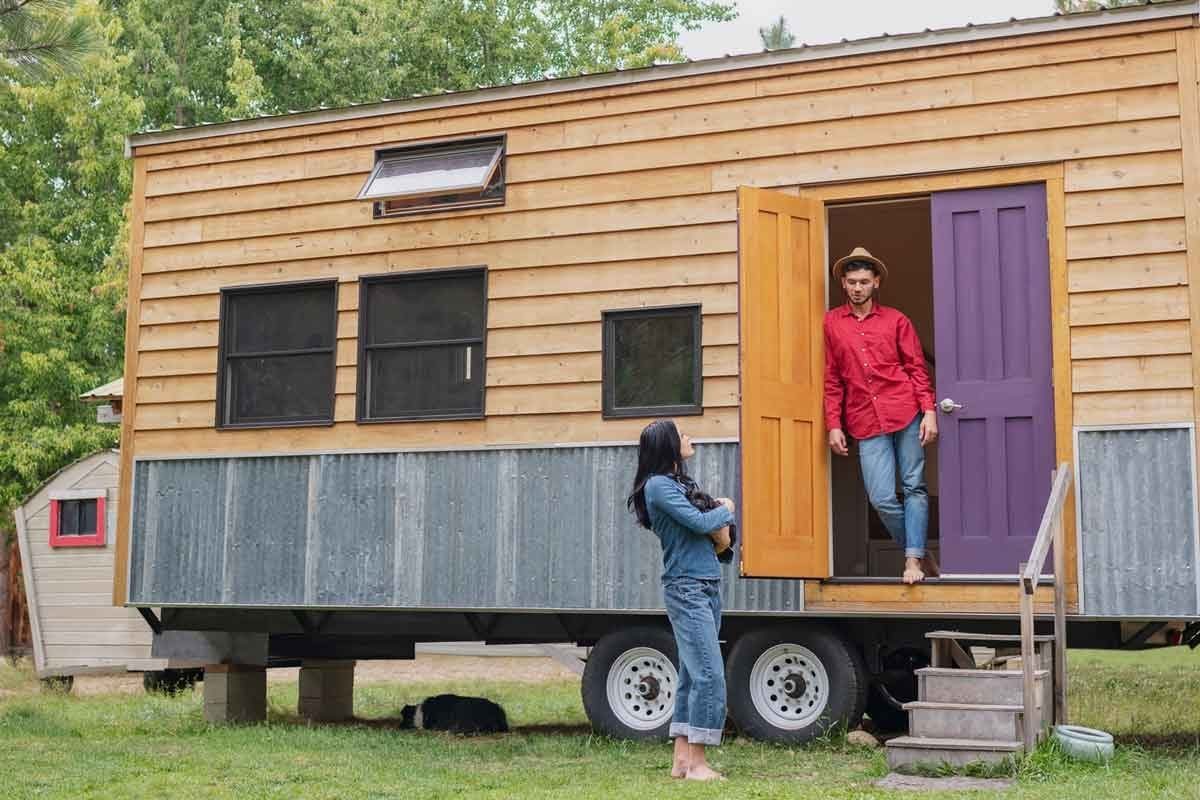 A man exits a tiny house with two colorful doors, while a woman stands nearby holding a cat in a green outdoor setting.