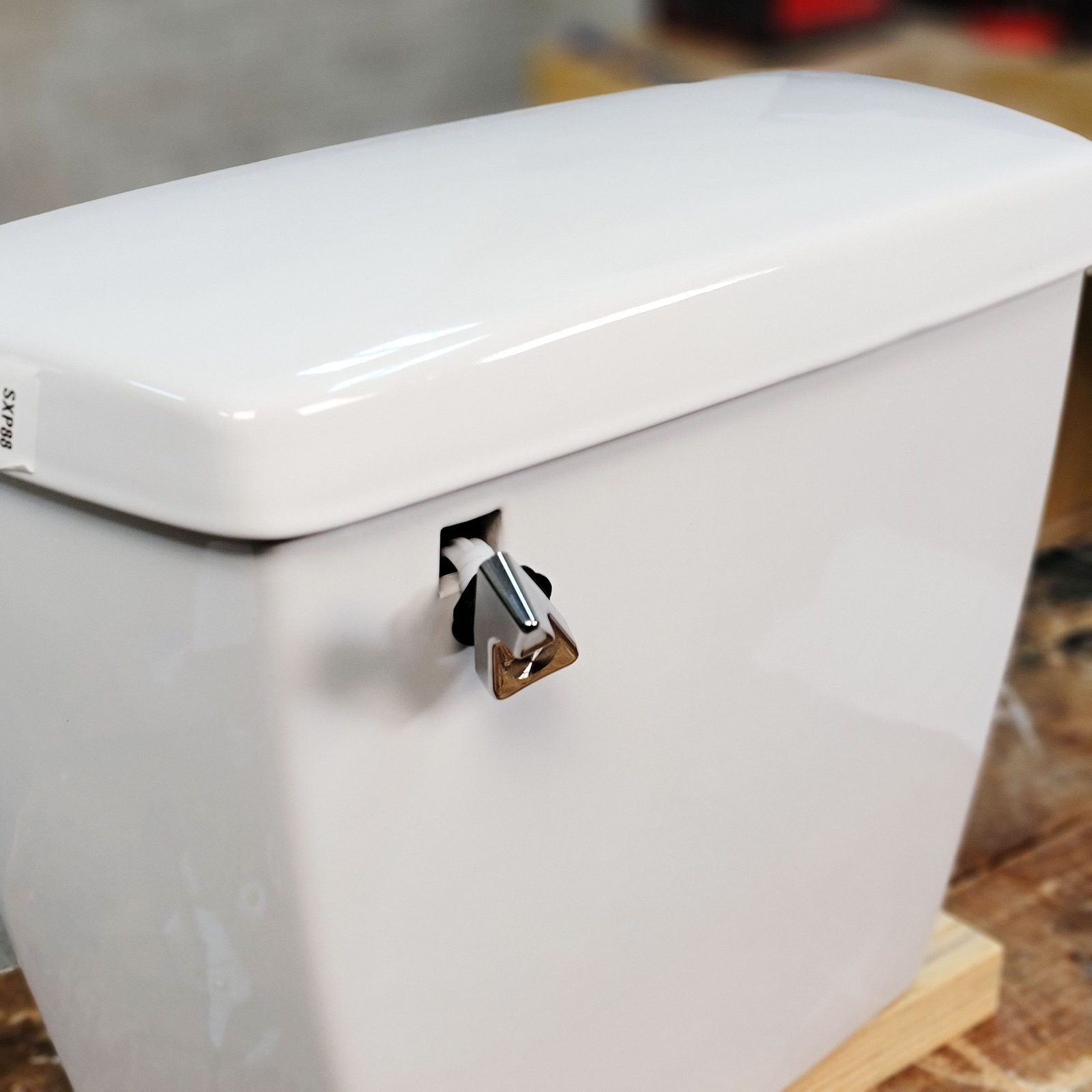A white toilet tank sits on a wooden surface, featuring a silver flush lever on the side, designed for easy operation in a bathroom setting.